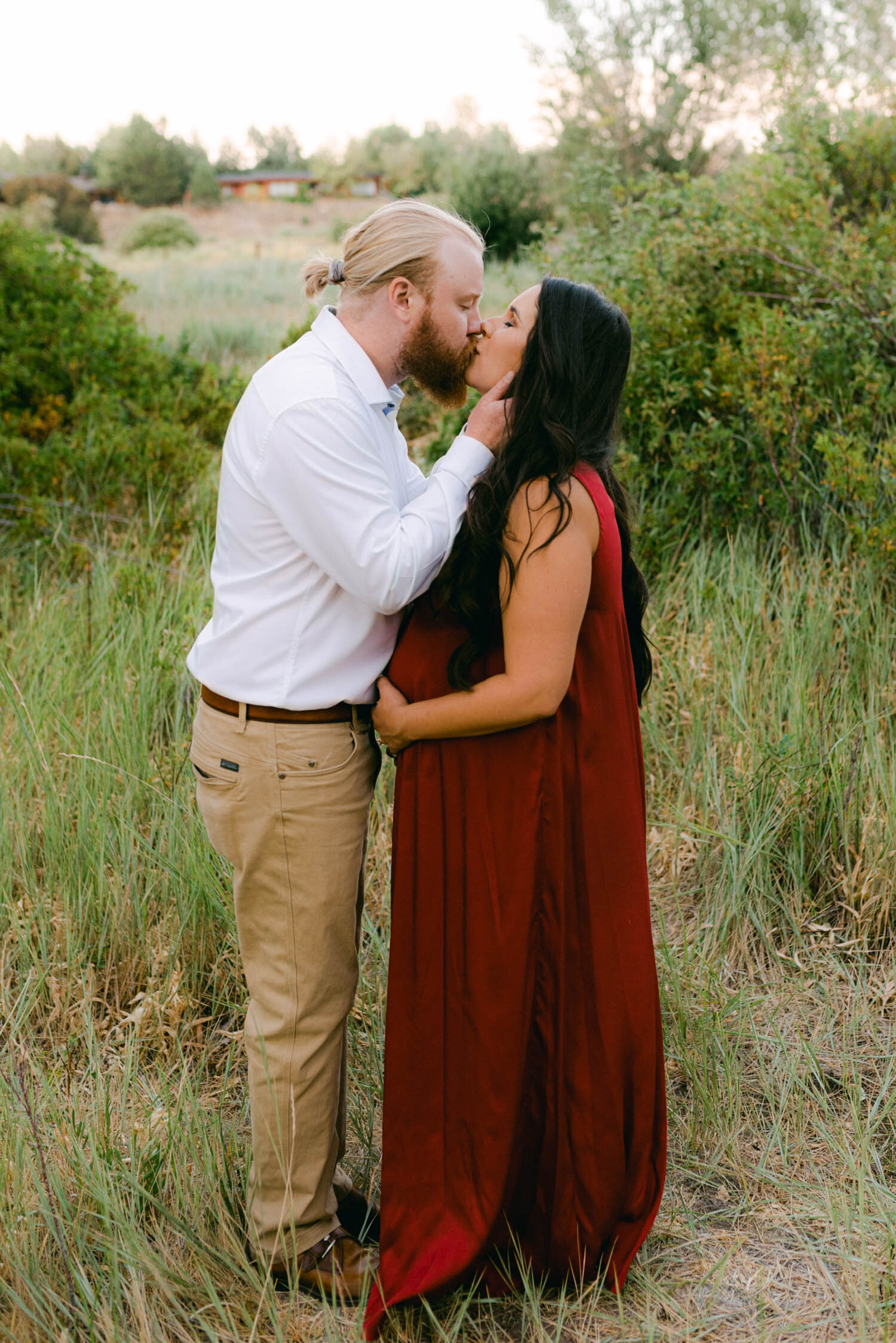 Golden hour maternity session, mom-to-be is wearing a red gown from rent the runway and kissing her partner. 