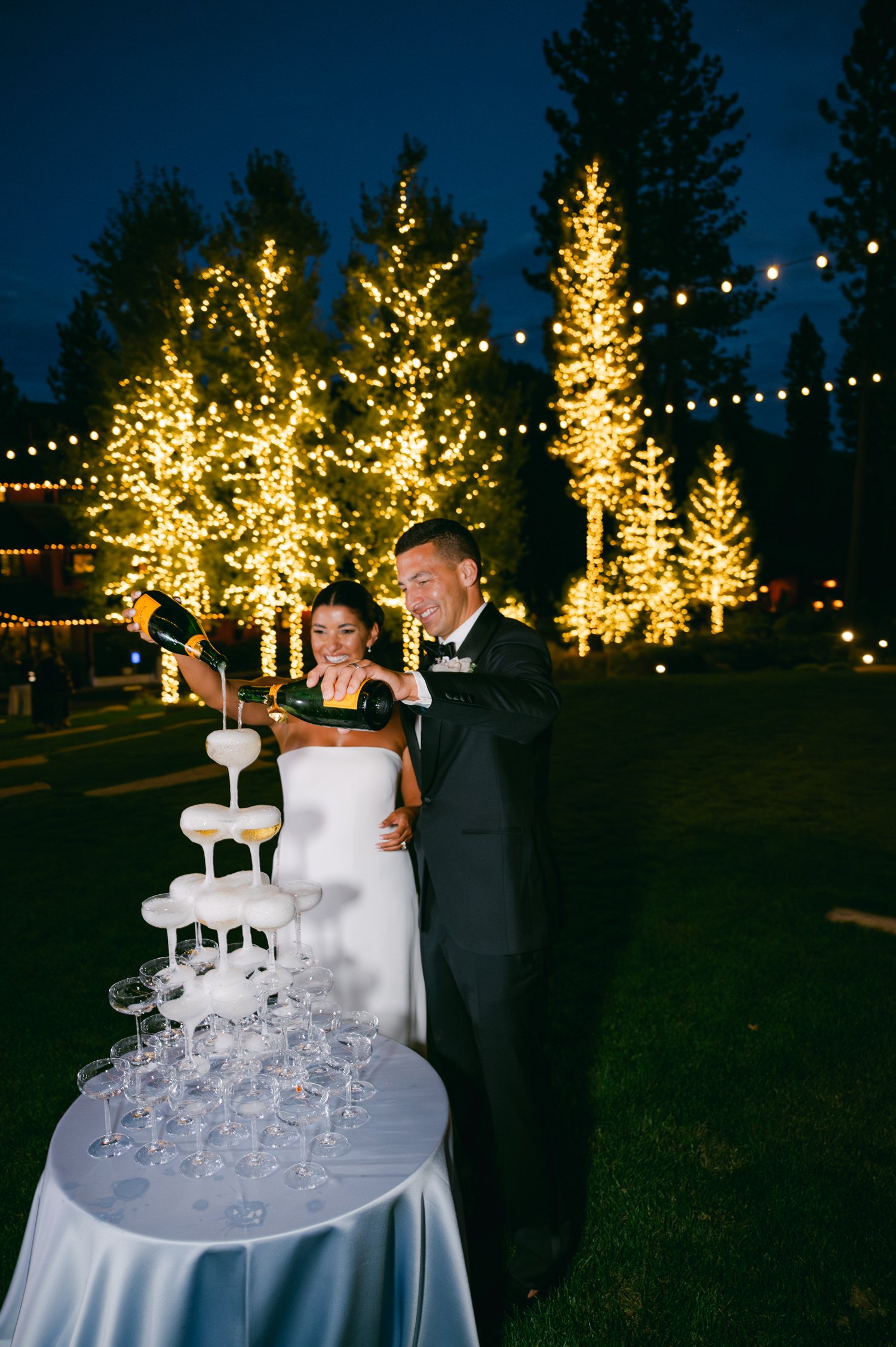 Martis Camp Wedding, photo of the newlywed couple pouring champagne with lit trees at the back 
