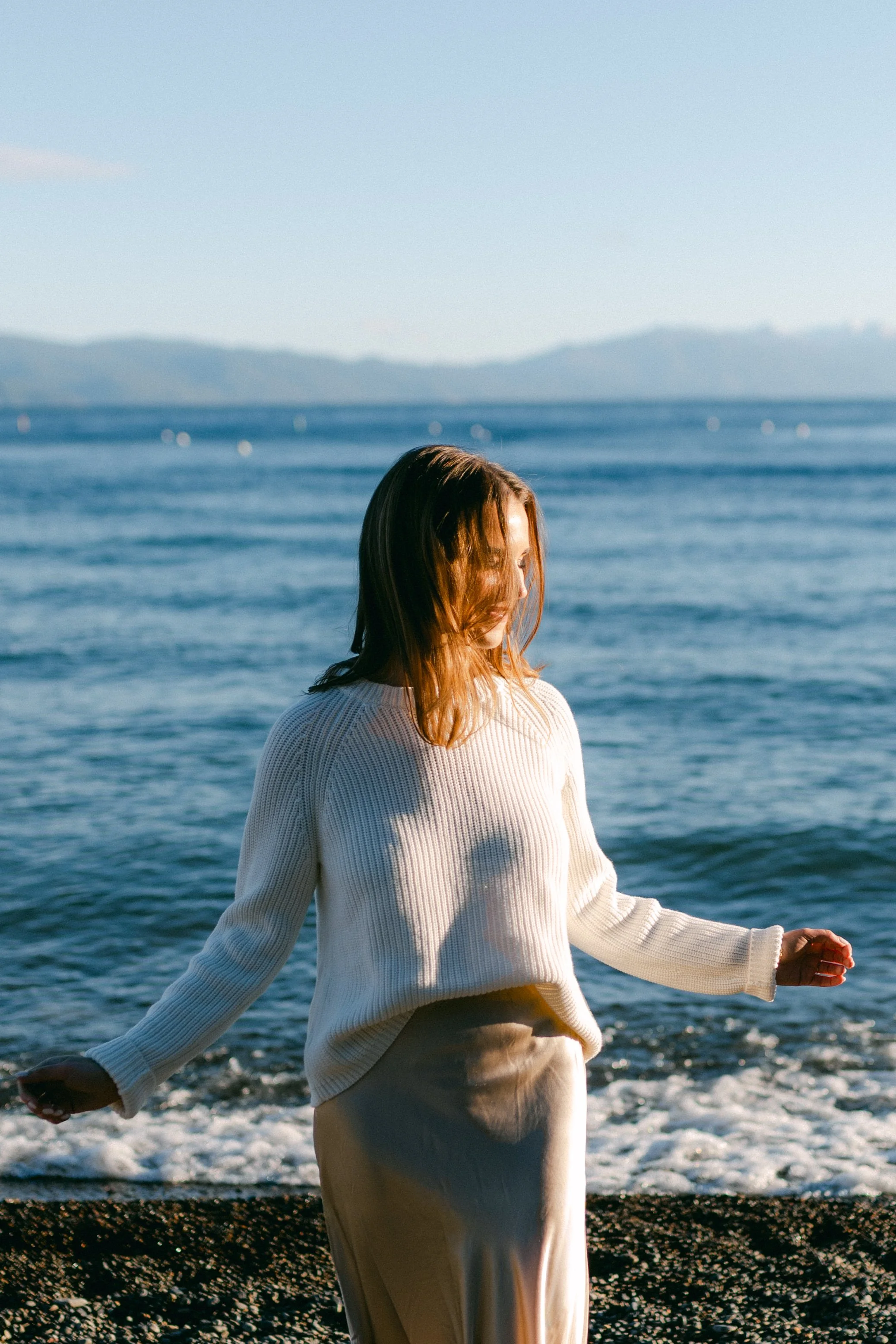Lake Tahoe Engagement session, photo of the bride wearing a white ribbed knit sweater and a neutral satin skirt while standing along the lake