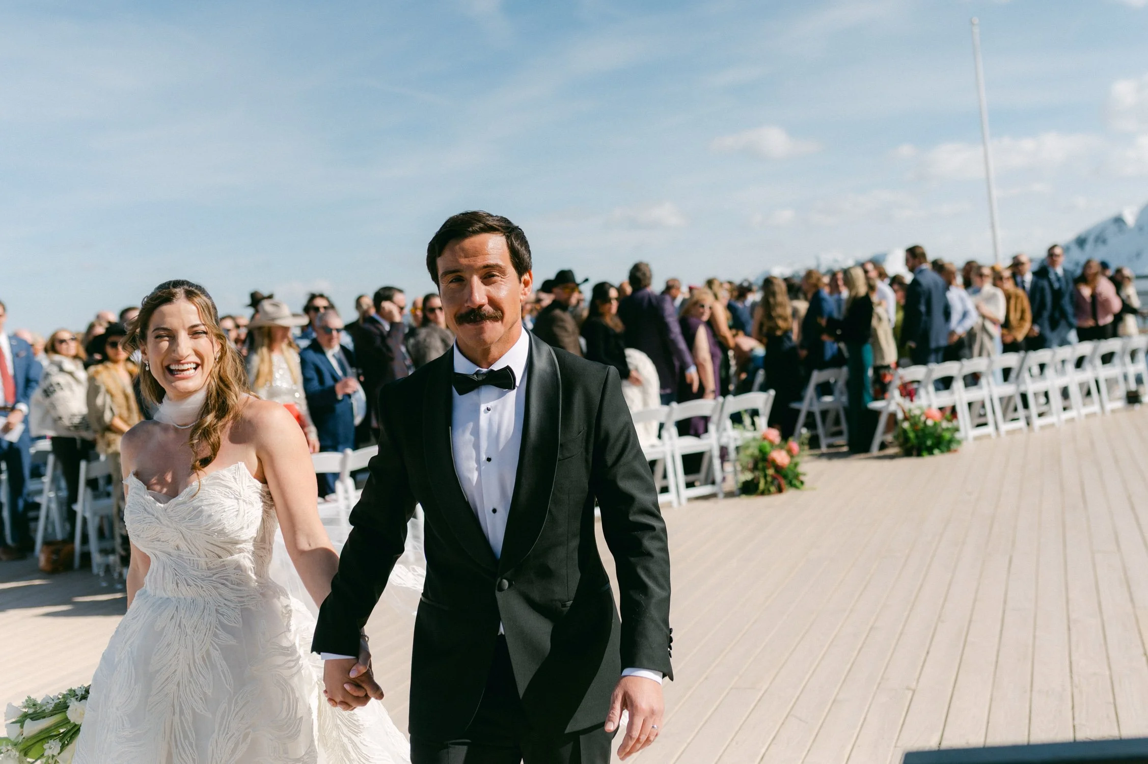 High Camp Ceremony, photo of the couple smiling and holding hands while walking down the aisle at an outdoor wedding ceremony. 