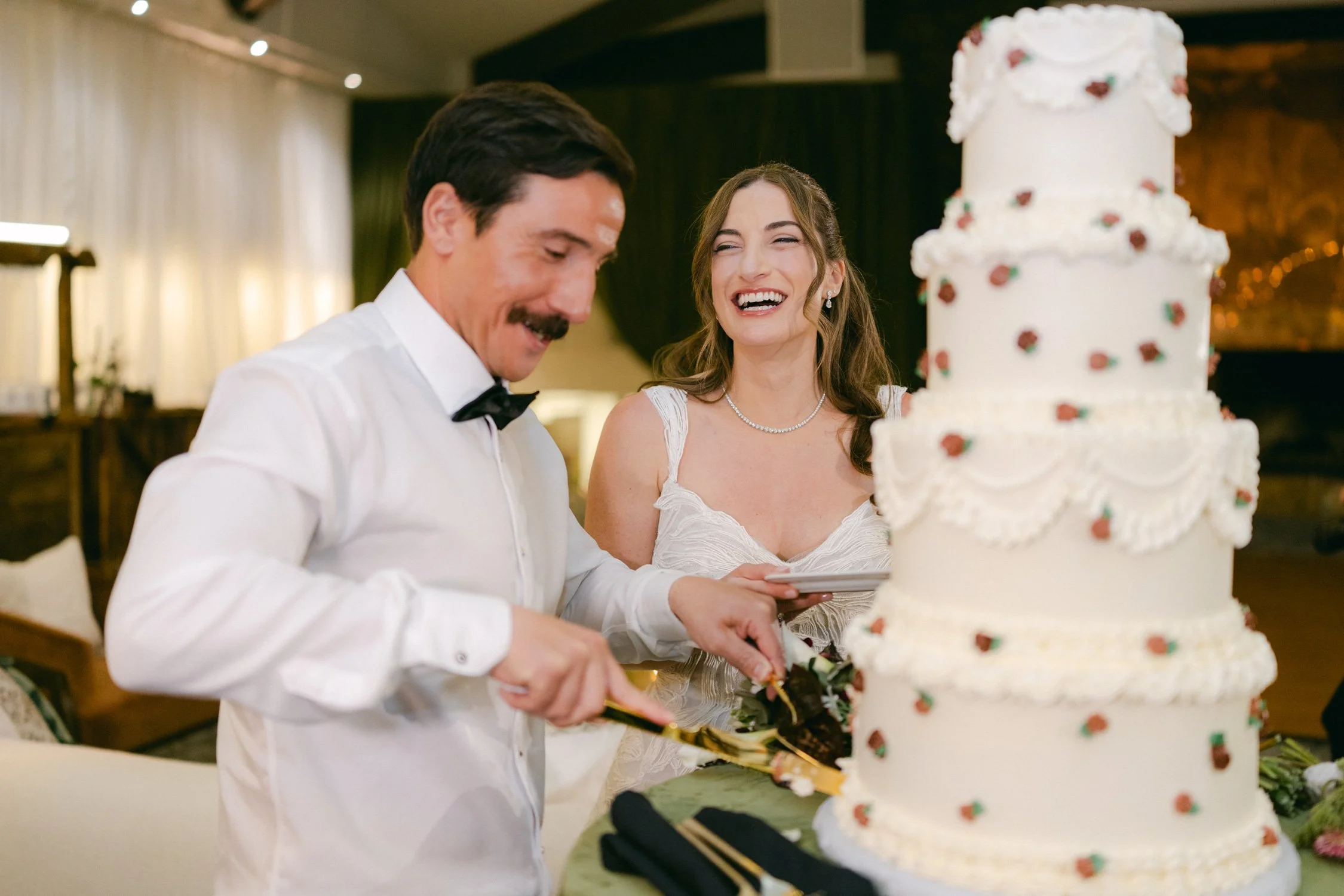 Palisades wedding venue, photo of a joyful moment from with the newlyweds featuring a custom multi-tiered cake decorated with delicate red rose accents