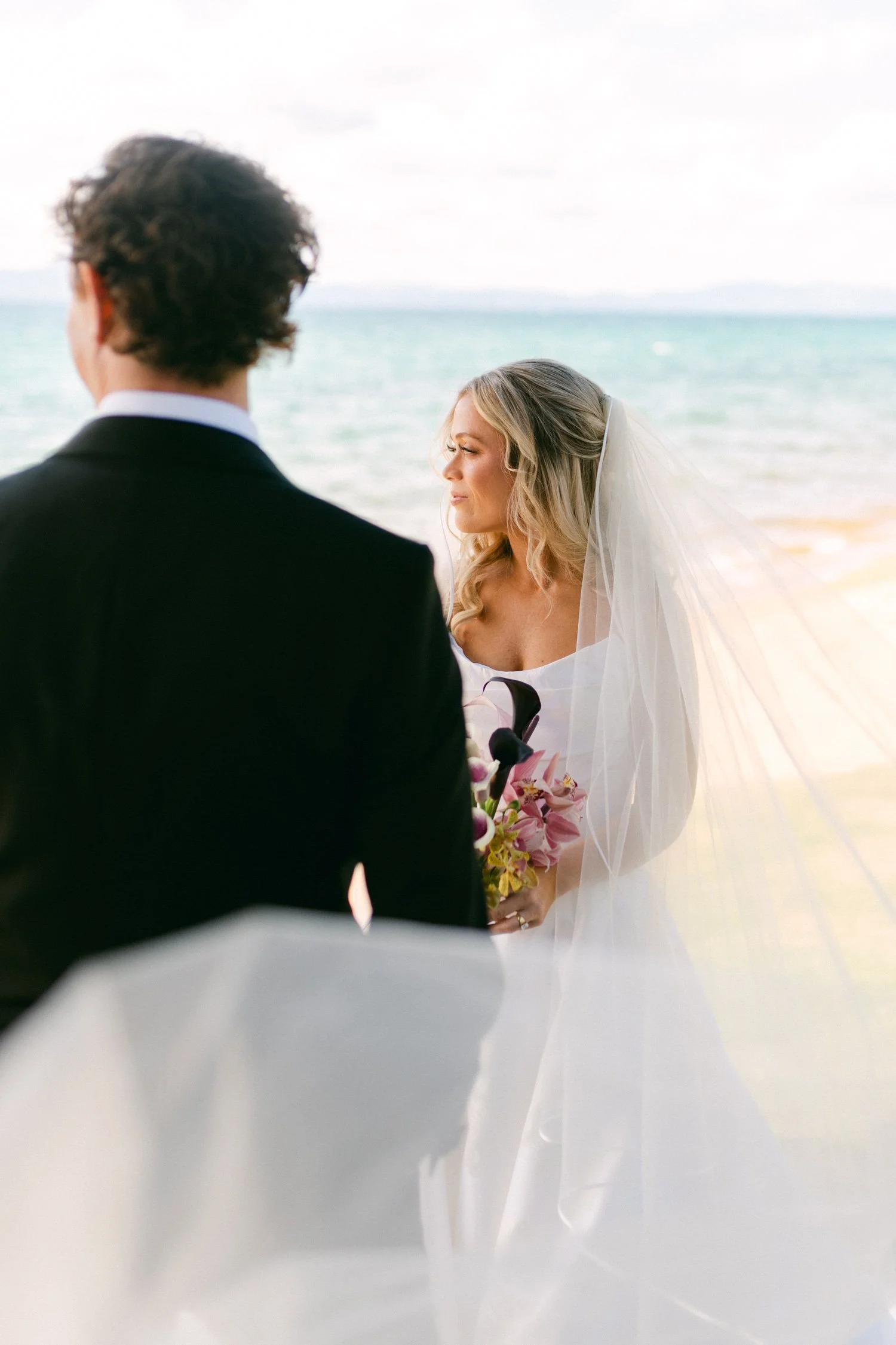 Edgewood maximalist wedding, photo of the bride in a simple, elegant wedding dress and a sheer veil, holding a bouquet of dark purple and pink flowers during a beach ceremony.