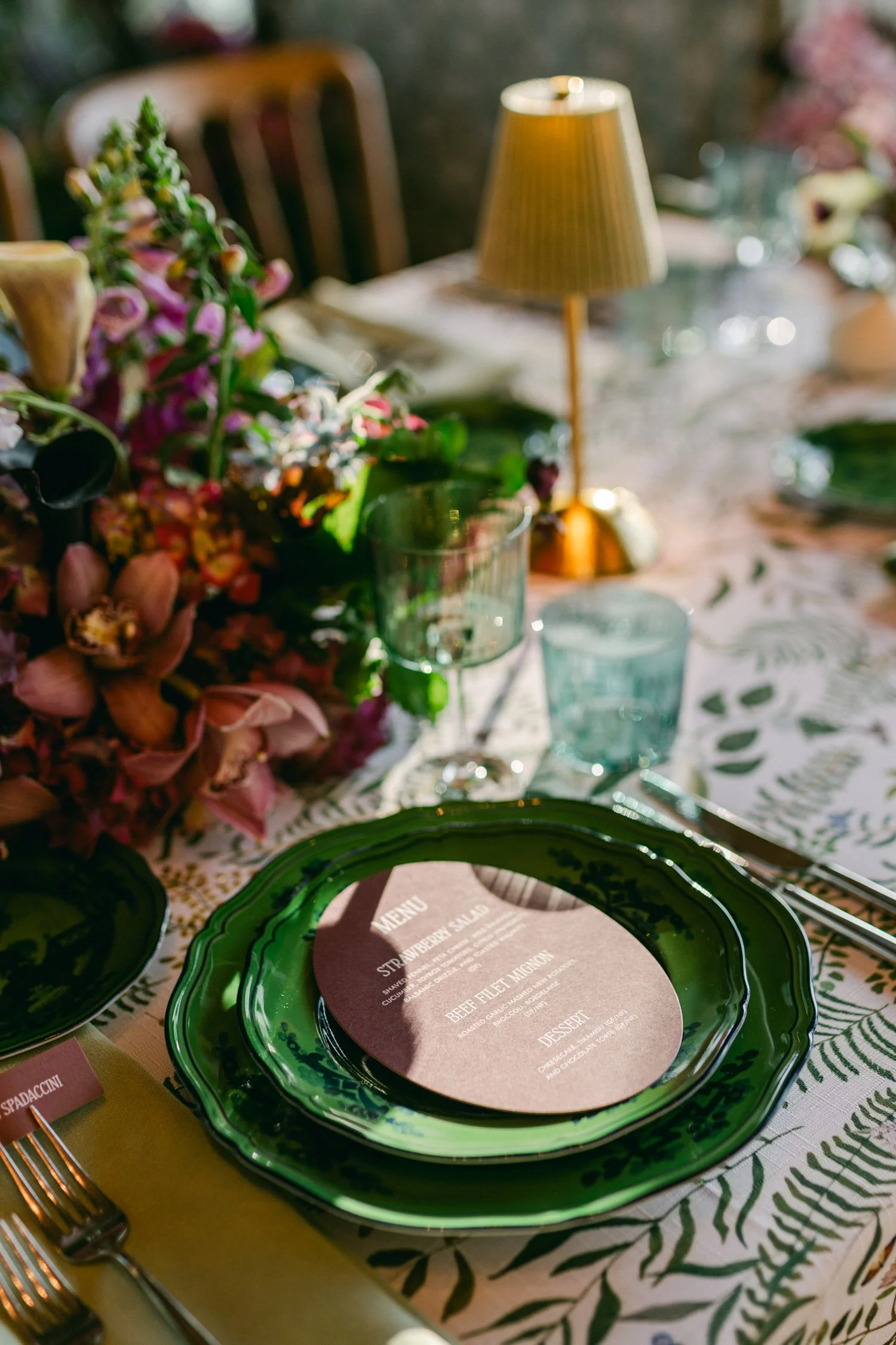 Edgewood maximalist wedding, photo of sophisticated wedding table setting featuring a botanical-print tablecloth, emerald green dinner plates, and a modern gold table lamp