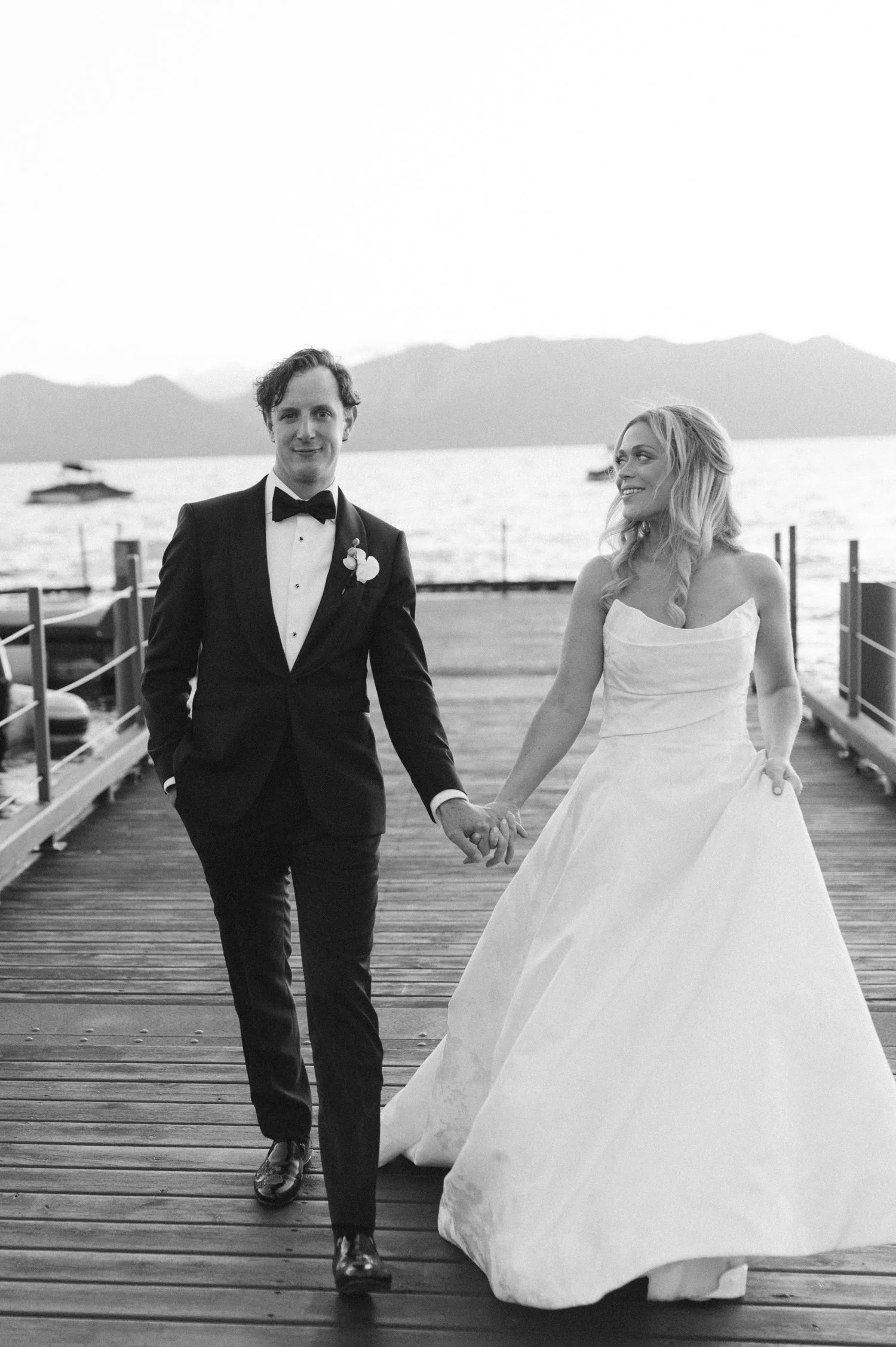 Edgewood maximalist wedding, photo of the newlyweds walking hand-in-hand along a wooden dock, set against a calm lake.