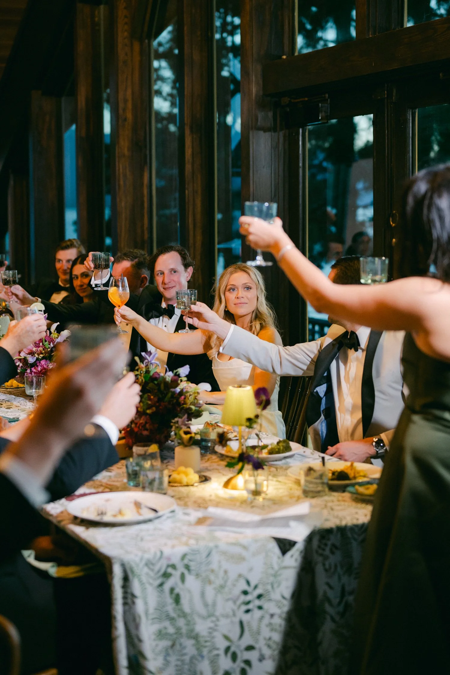Edgewood maximalist wedding, photo of an indoor wedding reception with guests raising glasses around a decorated table featuring floral arrangements and cocktail glasses