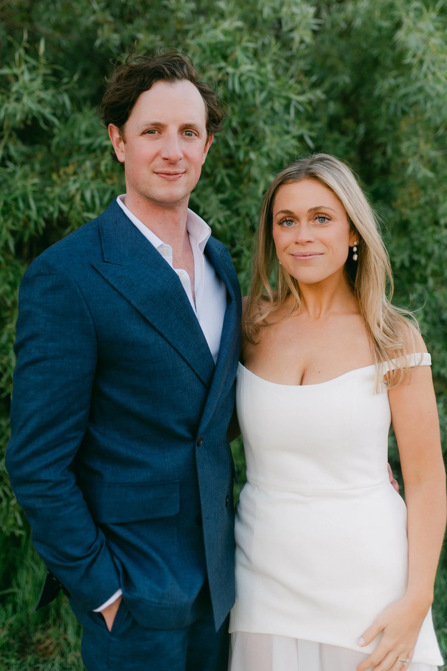 Riva Grill welcome party, photo of the couple posed against a lush green backdrop, featuring a tailored blue suit and a strapless white gown with a structured bodice