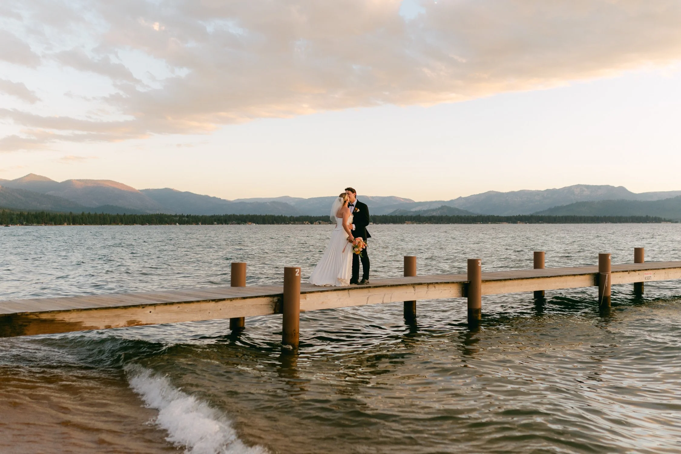 Lake Tahoe 4-H Camp wedding photos, photo of the newlyweds posing together during sunset on the dock with the stunning backdrop of Lake Tahoe and its surrounding mountains.
