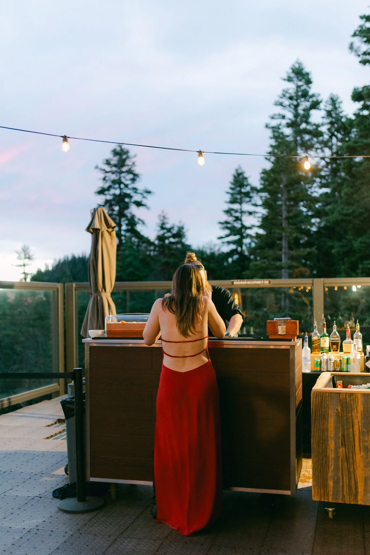 Zephyr Lodge wedding, photo of the wedding guest in a red gown with a bare back is ordering from an outdoor bar during a scenic sunset celebration.