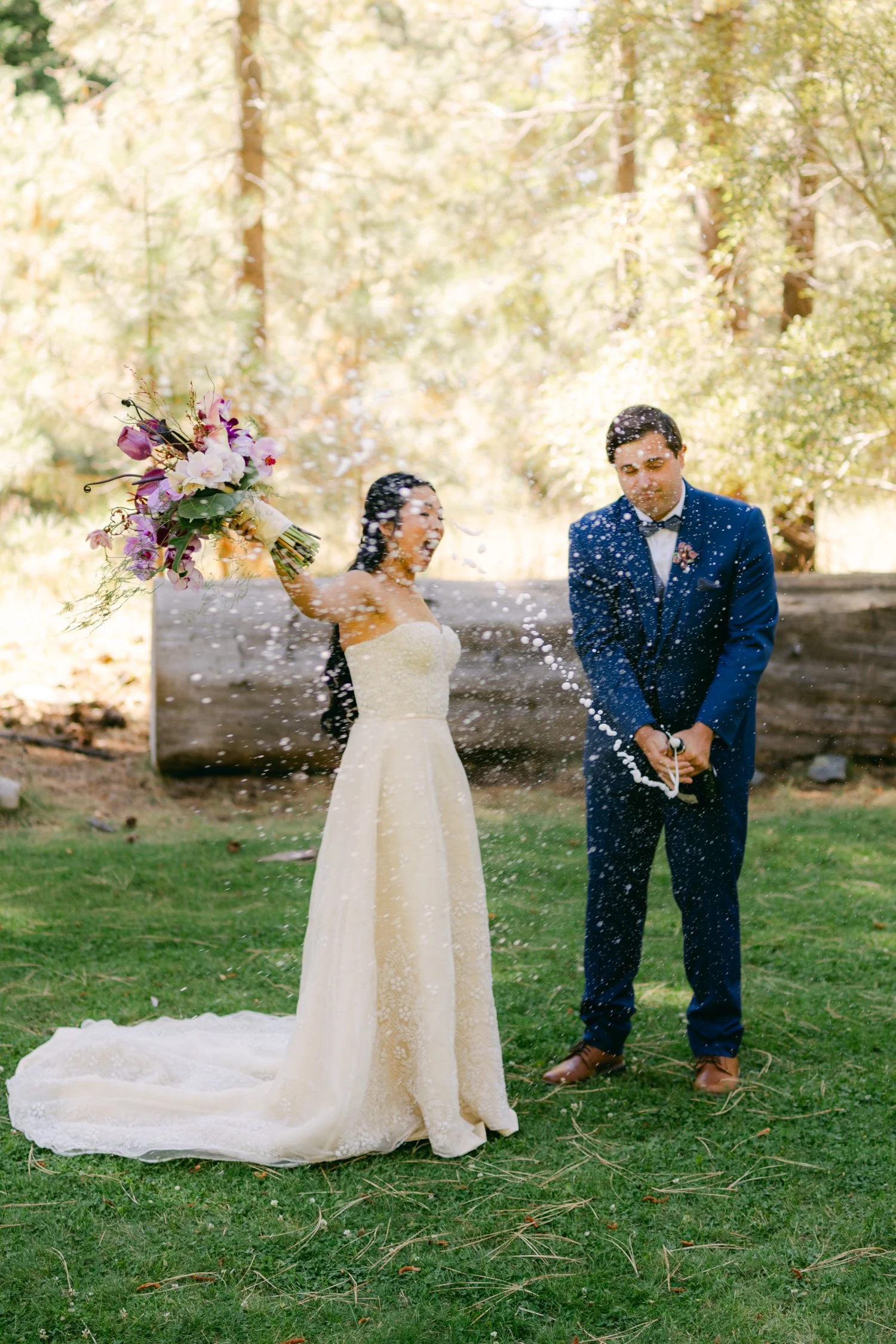 A Valhalla Lake Tahoe Wedding, photo of the bride in a strapless A-line wedding dress and a groom in a classic blue suit celebrating outdoors with a champagne pop.