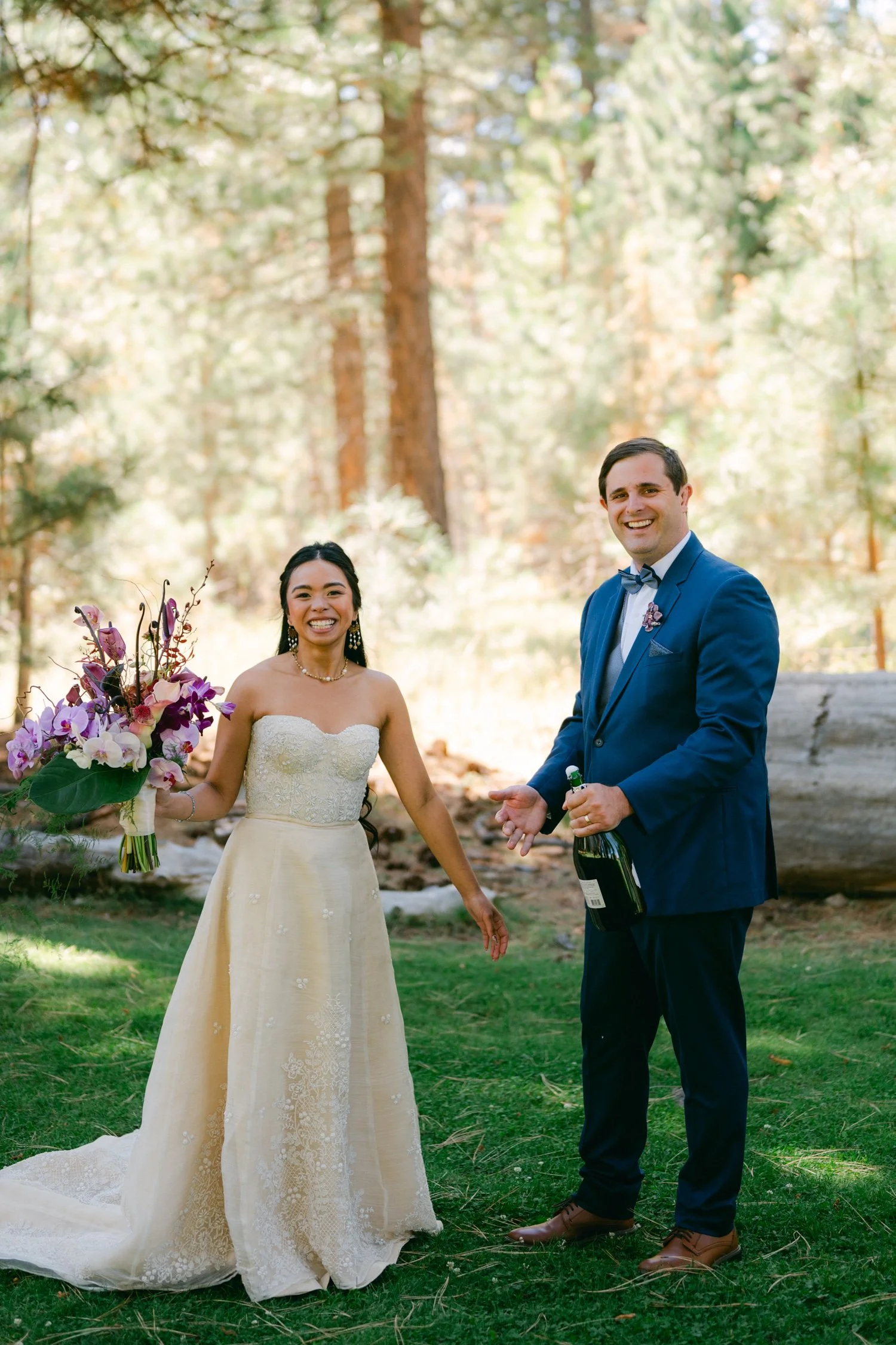 A Valhalla Lake Tahoe Wedding, photo of the couple posing outdoors in a wooded area, with the bride holding a vibrant floral bouquet and the groom holding a champagne bottle.