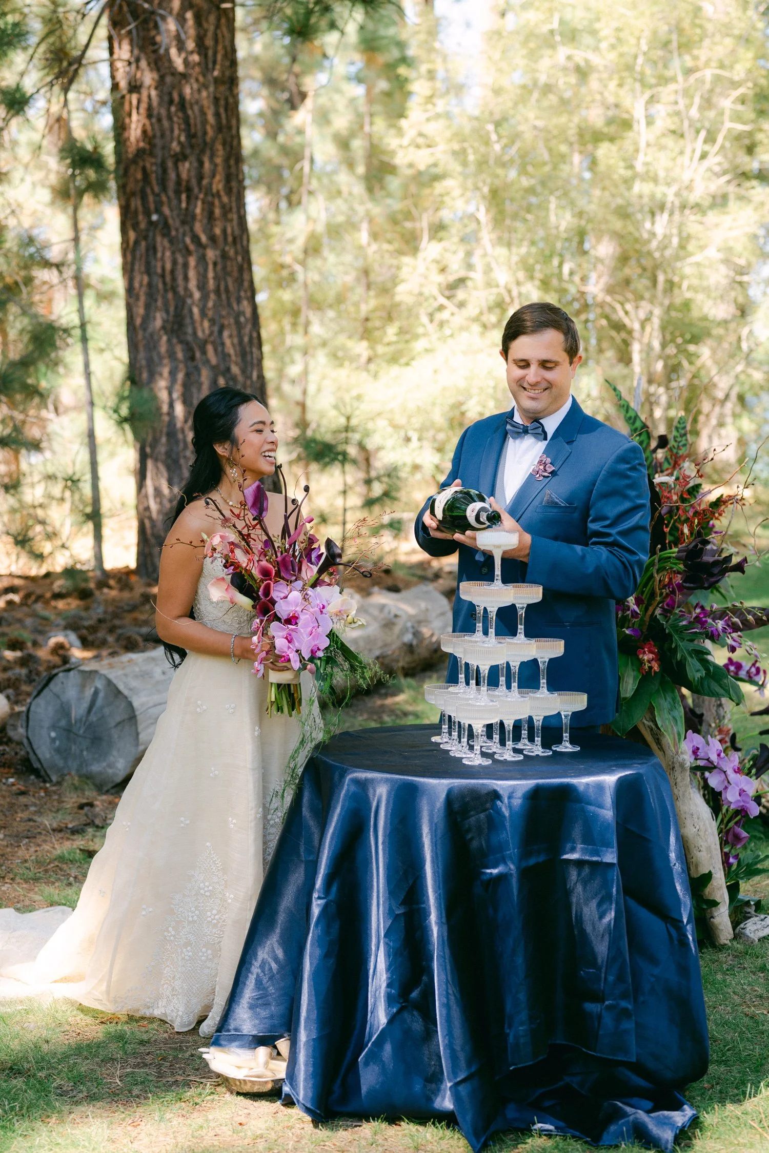 A Valhalla Lake Tahoe Wedding, photo of the bride and groom pouring champagne over a tiered champagne tower set up on a table draped with blue satin fabric.