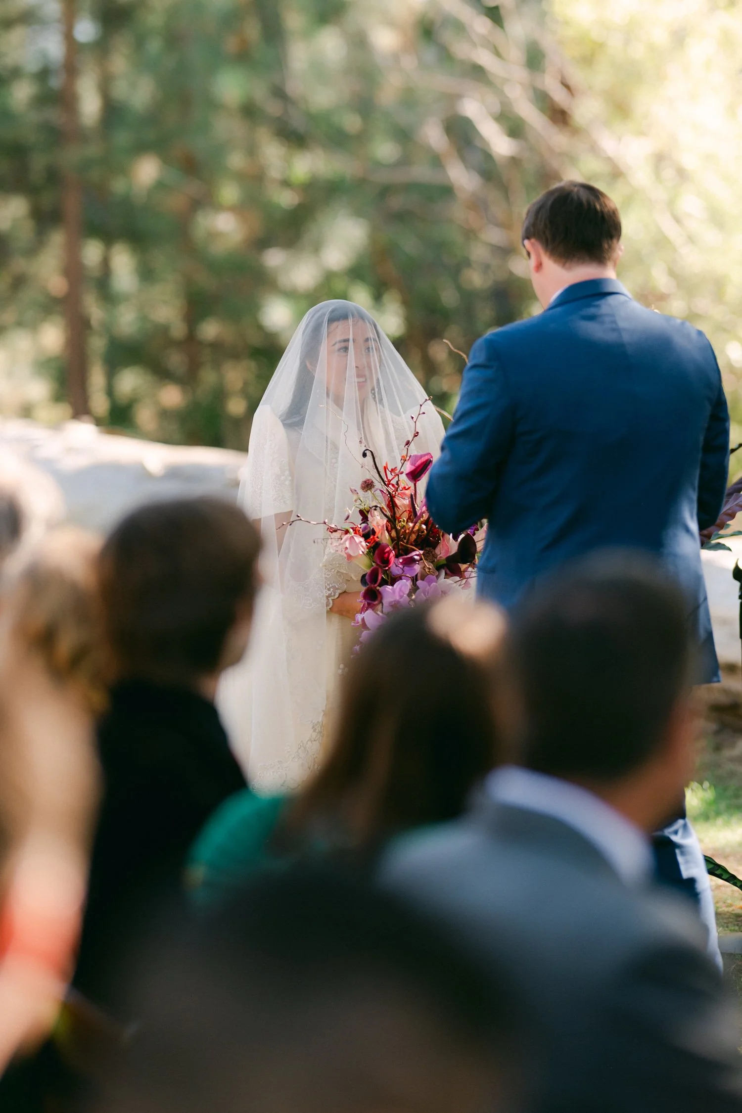A Valhalla Lake Tahoe Wedding, photo of an outdoor wedding ceremony in a wooded area, with a bride wearing a veil and holding a colorful bouquet while facing the groom in a blue suit.