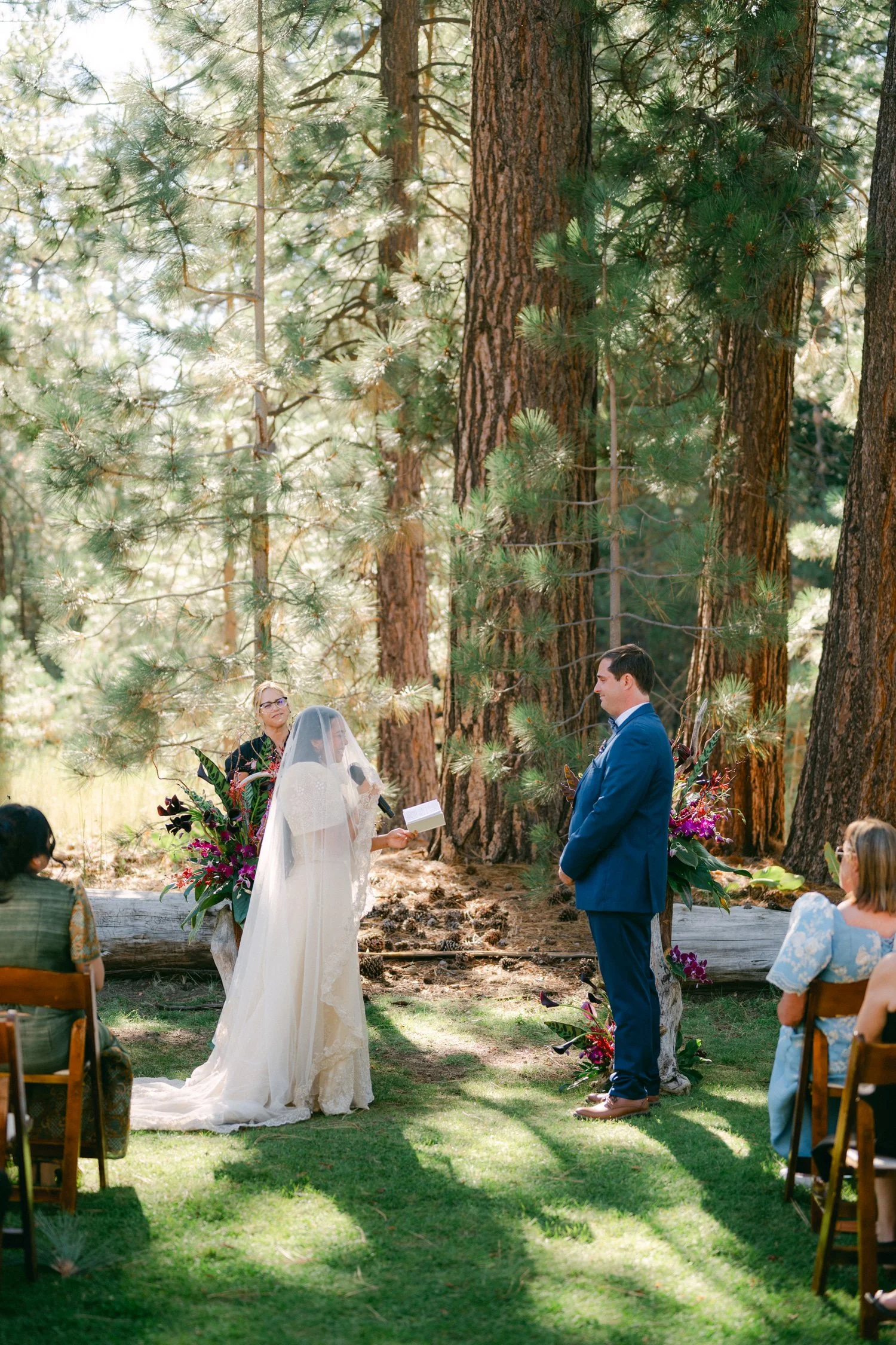 A Valhalla Lake Tahoe Wedding, photo of an outdoor forest wedding ceremony among tall pine trees with the bride wearing a flowing, light-colored gown with a veil or shawl over her head