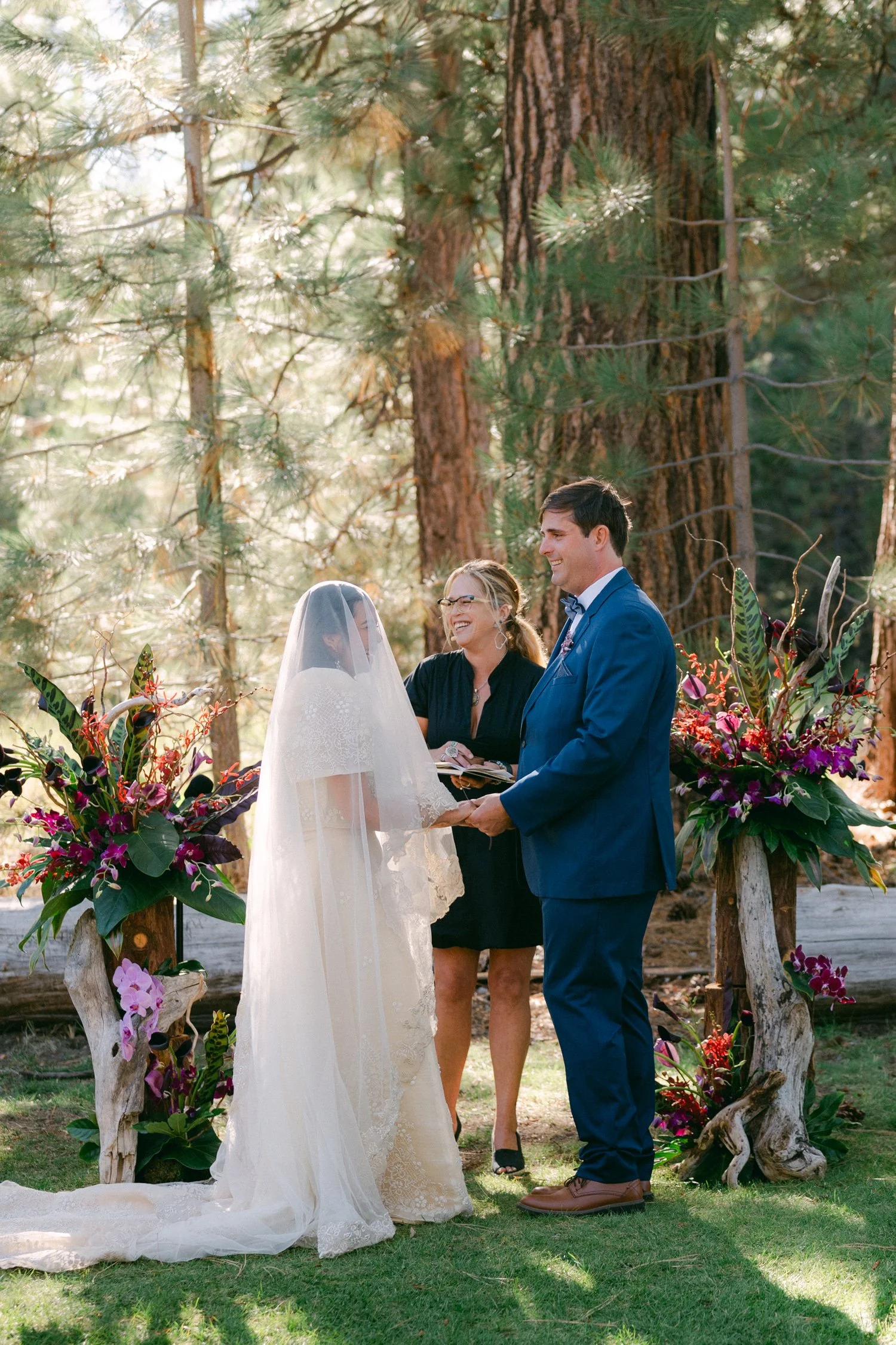 A Valhalla Lake Tahoe Wedding, photo of an outdoor wedding ceremony taking place in a pine forest, featuring the bride in a veil and cape and the groom in a blue suit