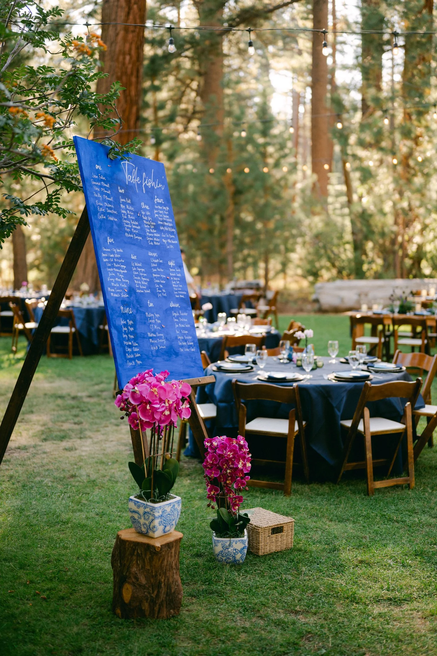 A Valhalla Lake Tahoe Wedding, photo of outdoor woodland wedding reception featuring a blue wooden seating chart and round tables set on a grassy lawn