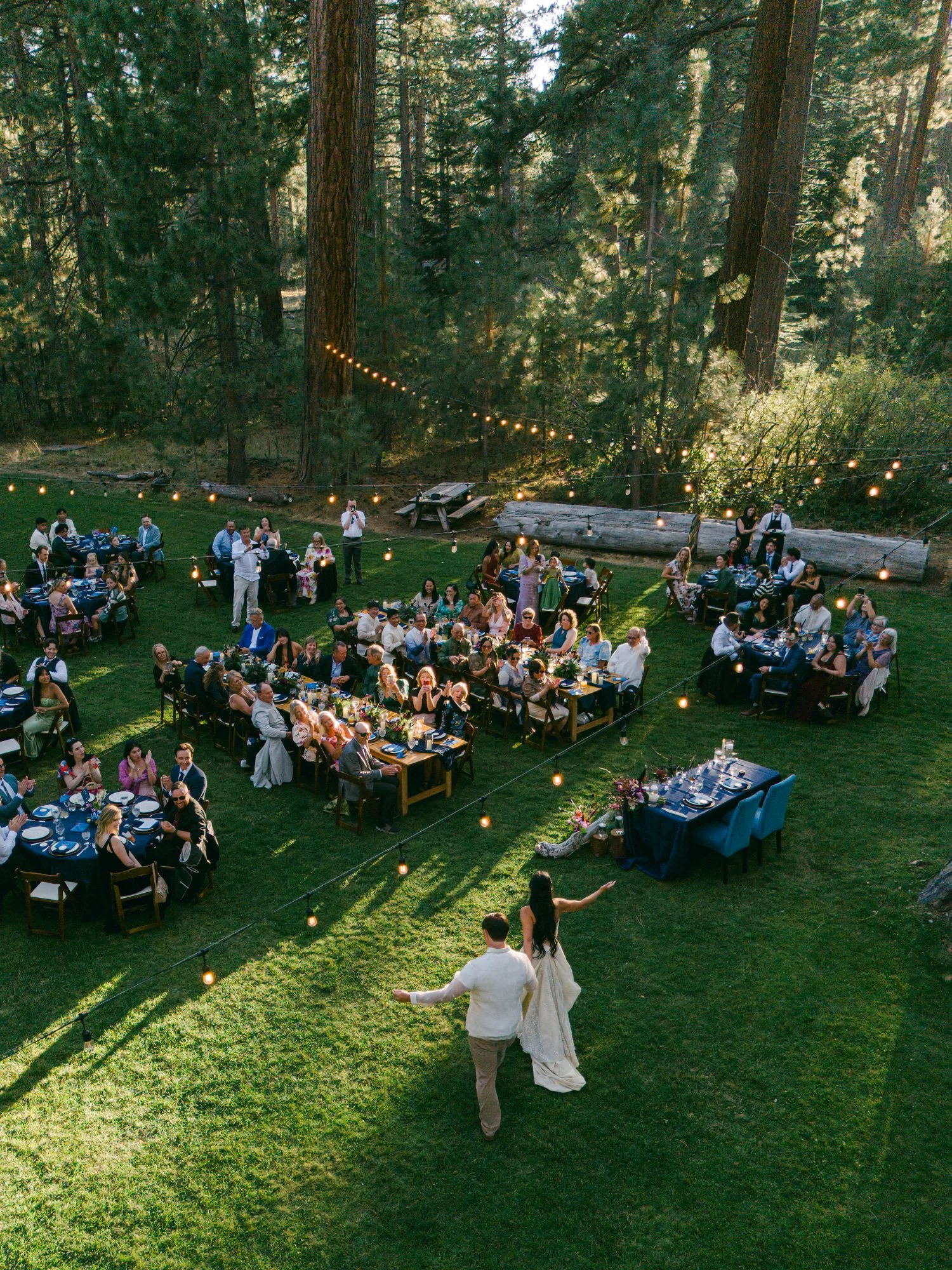 A Valhalla Lake Tahoe Wedding, photo of  outdoor forest wedding reception with guests are seated at long, candlelit tables arranged on a lush lawn surrounded by tall pine trees