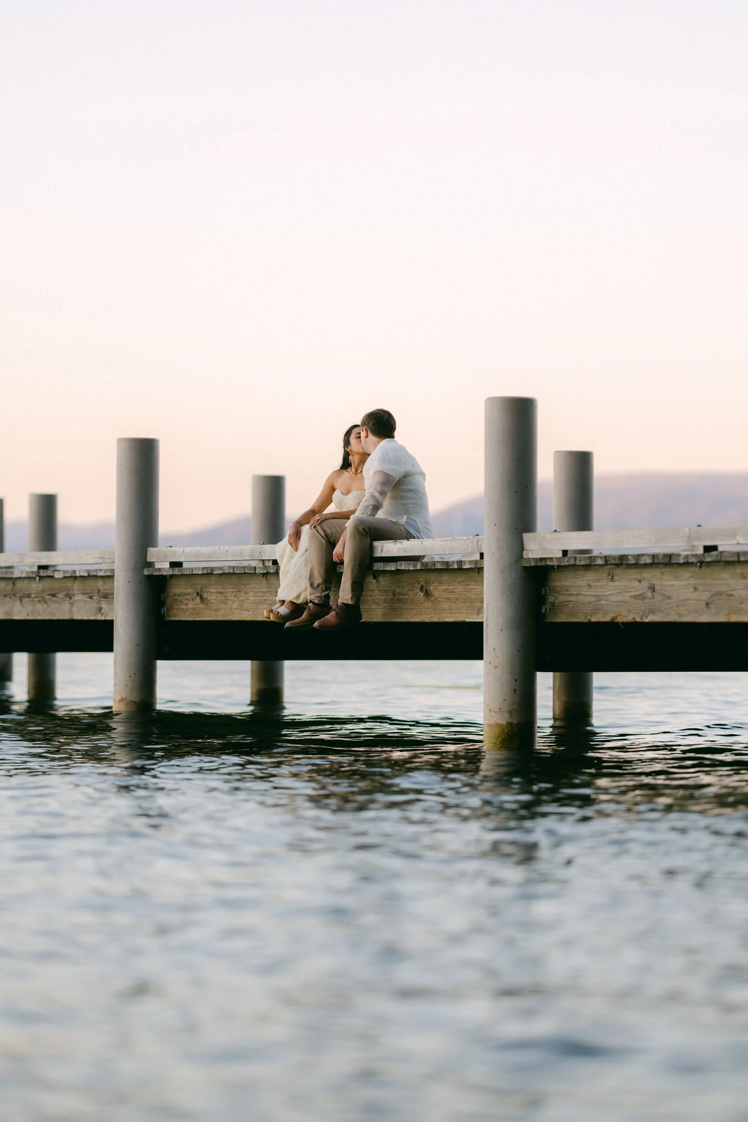 A Valhalla Lake Tahoe Wedding, photo of the couple sitting and kissing on a wooden pier at Lake Tahoe, framed against calm water and a soft sunset
