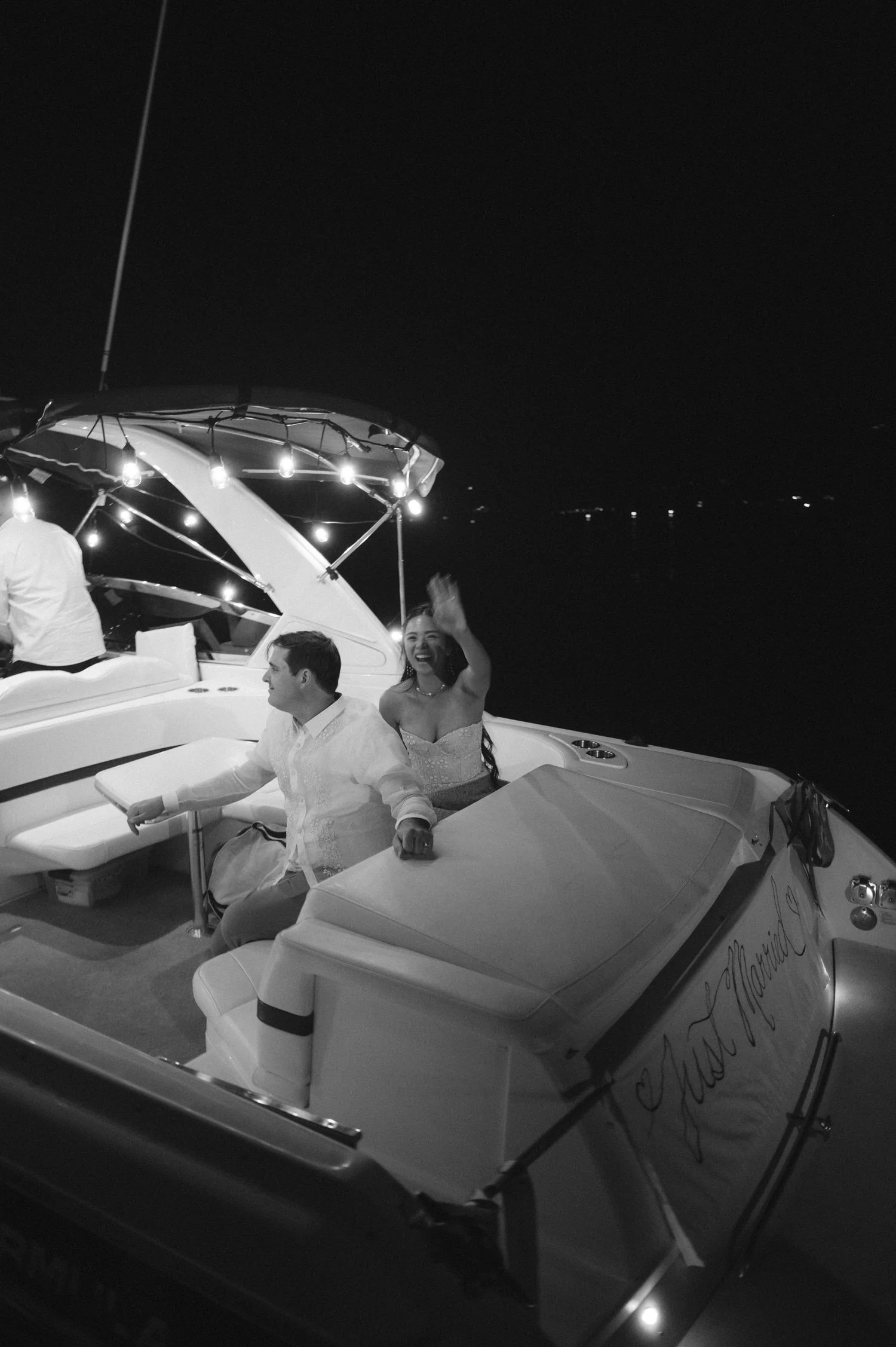 A Valhalla Lake Tahoe Wedding, photo of the newlywed couple waving from the stern of a boat decorated with a "Just Married" sign at night.
