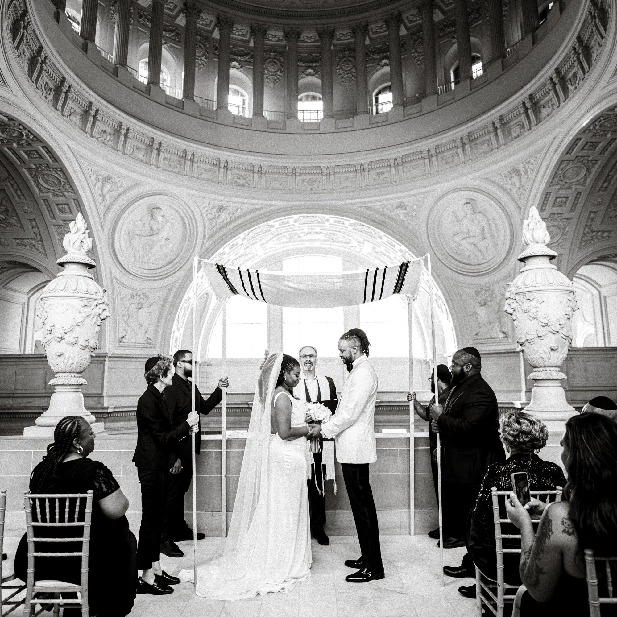 A bride and groom stand under a chuppah on the fourth floor, captured by an SF City Hall wedding photographer in an ornate hall.