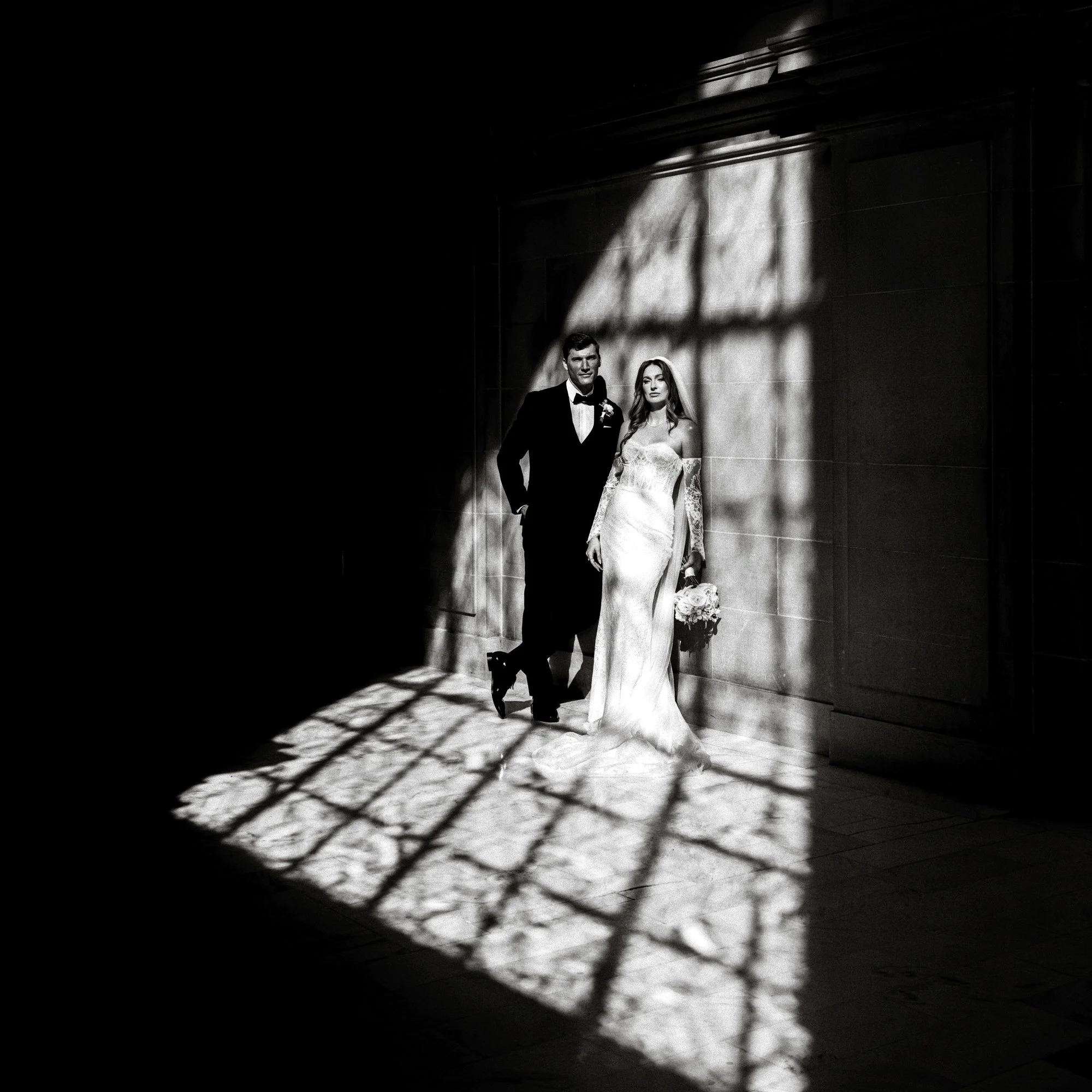 A bride and groom stand together in sunlit San Francisco City Hall, surrounded by shadow, in black and white editorial photography.