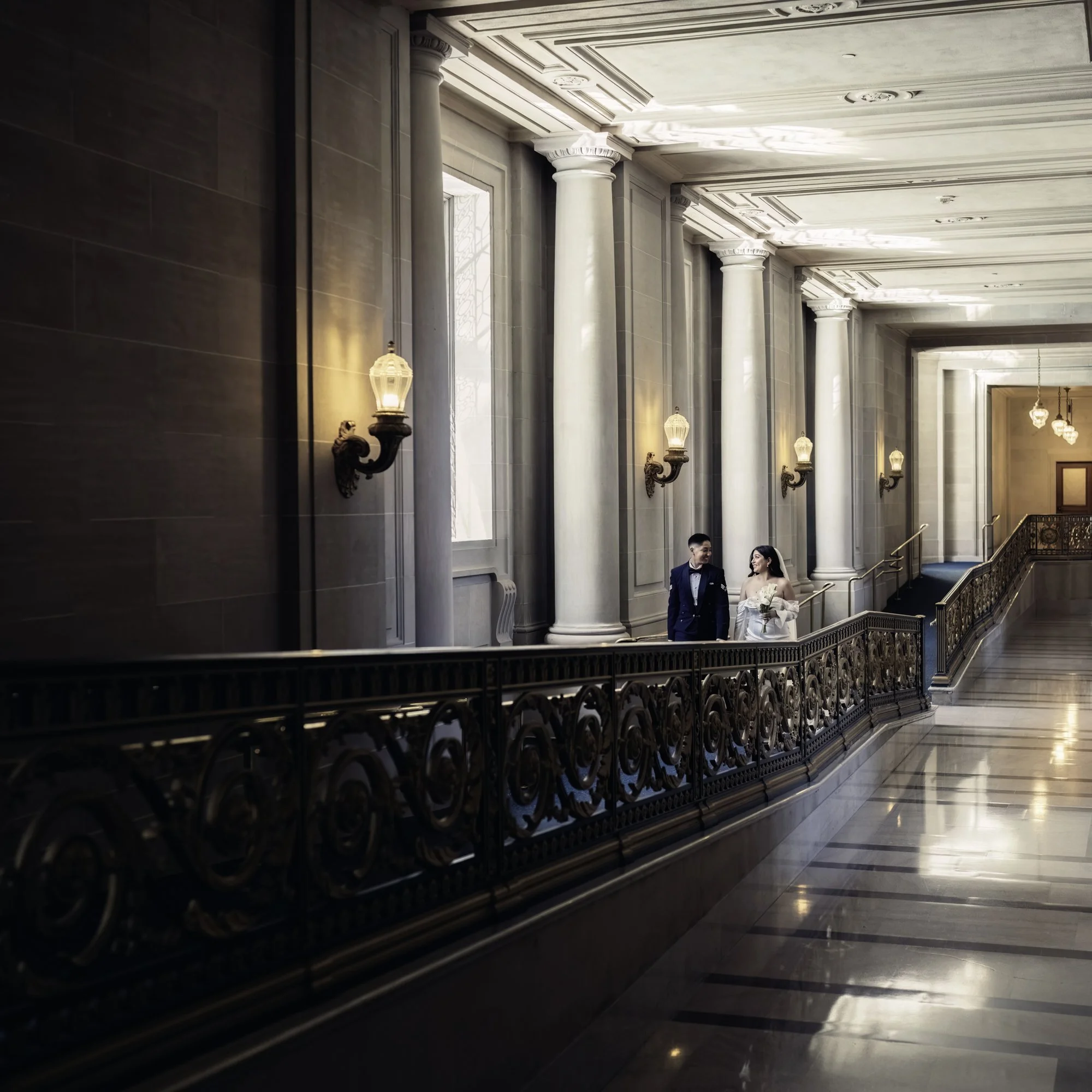 A couple celebrates their LGBTQ wedding, walking down grand SF City Hall halls with tall columns and sunlit windows.