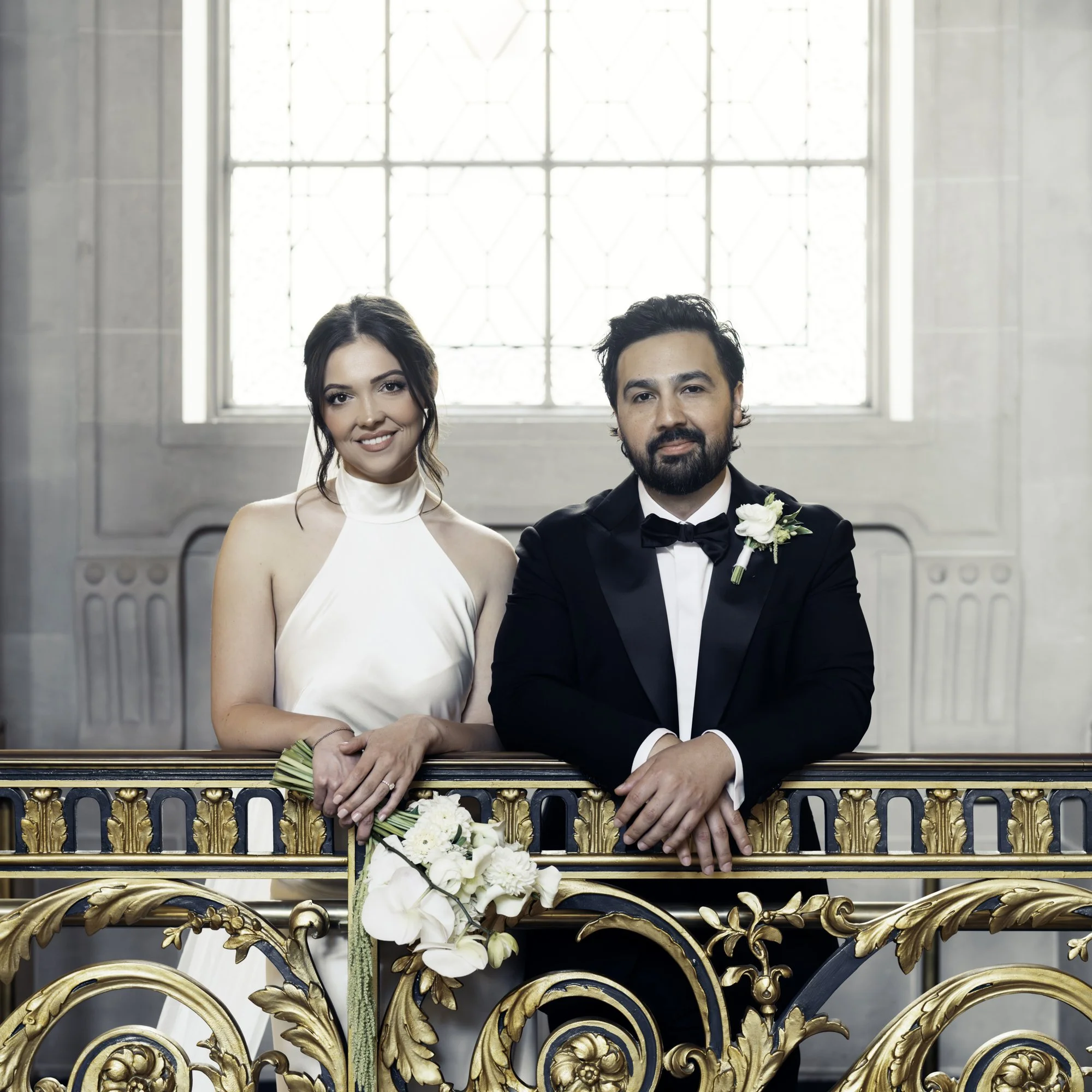 Bride in white and groom in tuxedo smile by ornate gold railing—captured by a San Francisco City Hall wedding photographer.