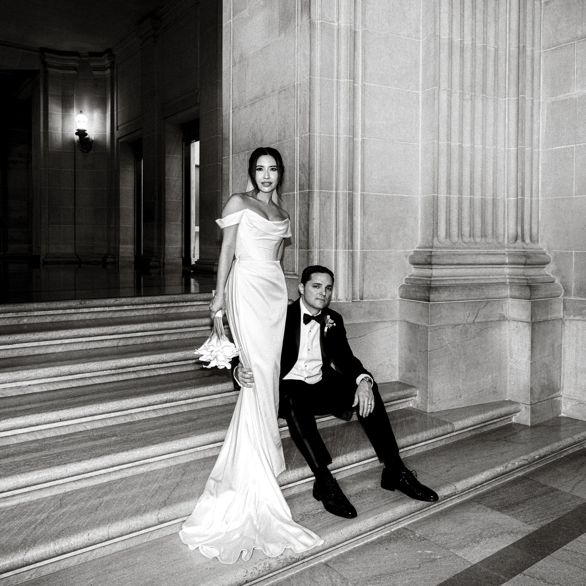 A bride in an elegant gown stands with her groom on marble steps, captured by a San Francisco City Hall wedding photographer.