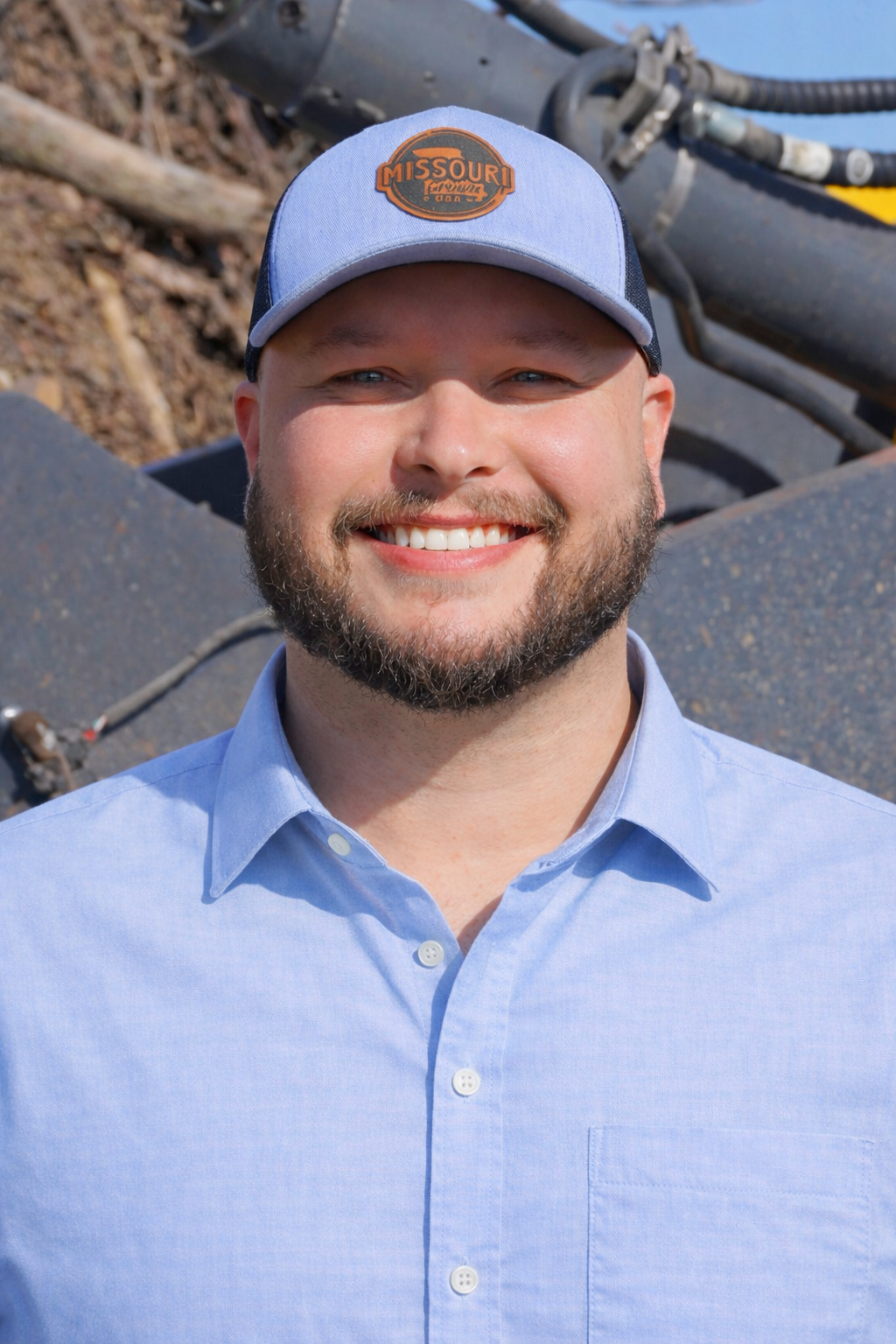 A man smiling outdoors in front of a piece of heavy machinery with dirt and debris. He is wearing a gray cap with an orange logo and a black vest over a white shirt.