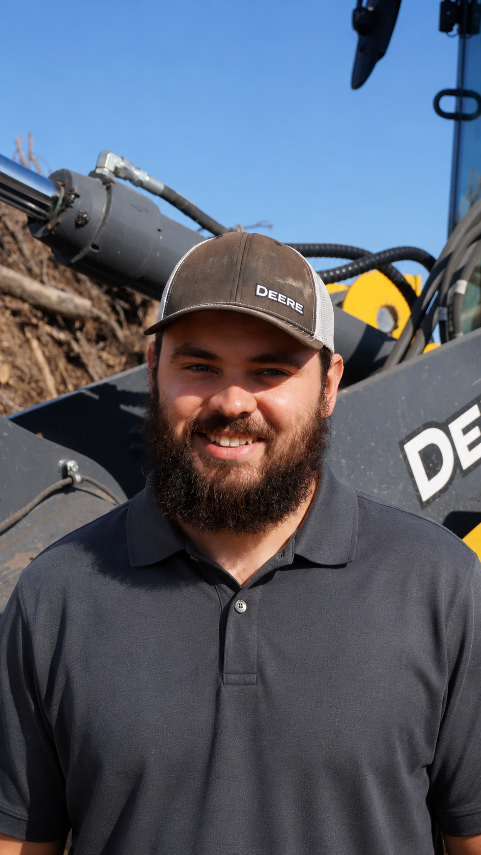A smiling man with a beard wearing a gray Deere baseball cap and a red hoodie with yellow graphics and text, standing outdoors near a construction or agricultural machine.