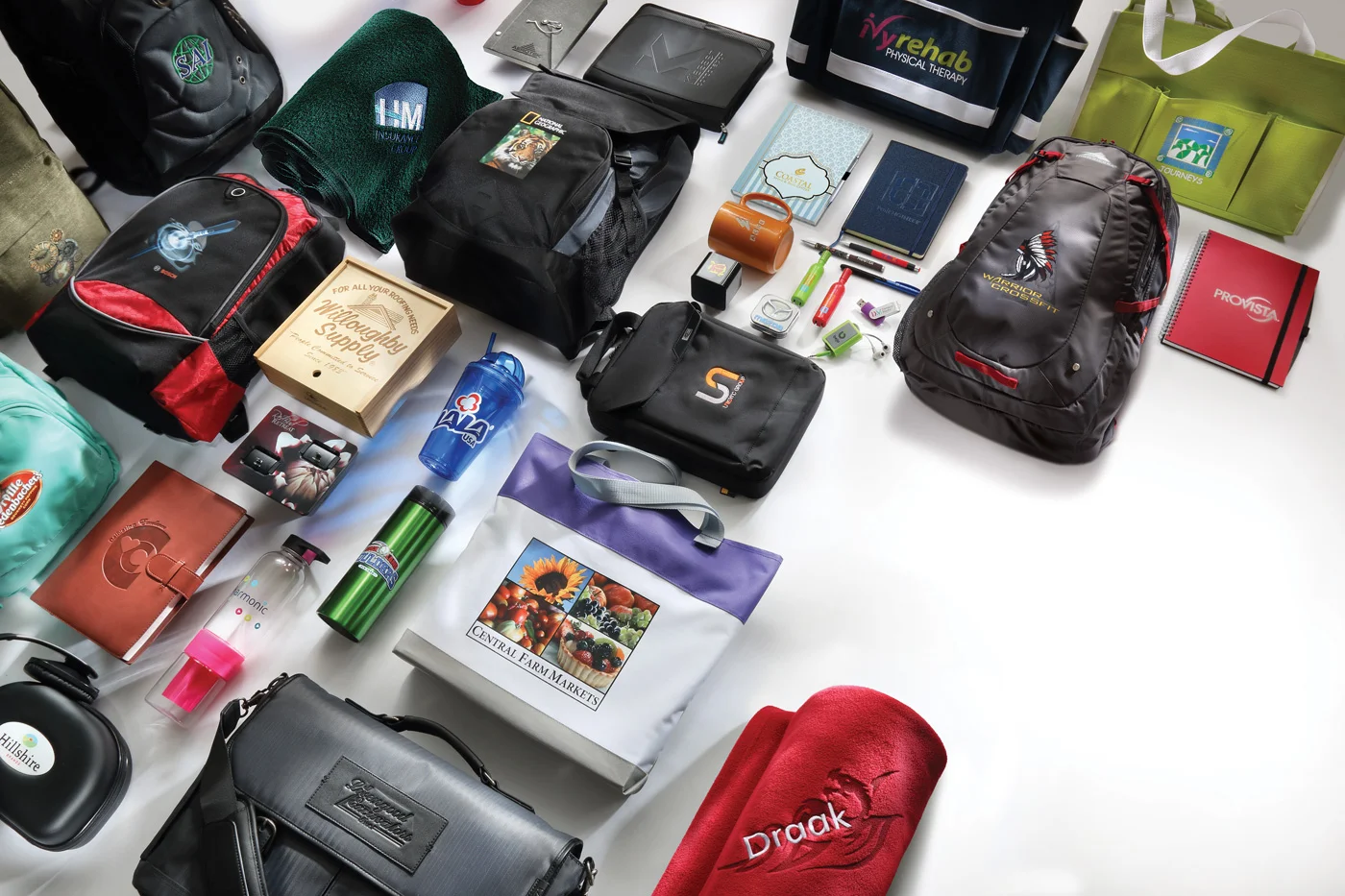 Various travel and outdoor gear, including backpacks, notebooks, water bottles, a fleece blanket, a tote bag, and personal items, are laid out on a white surface.