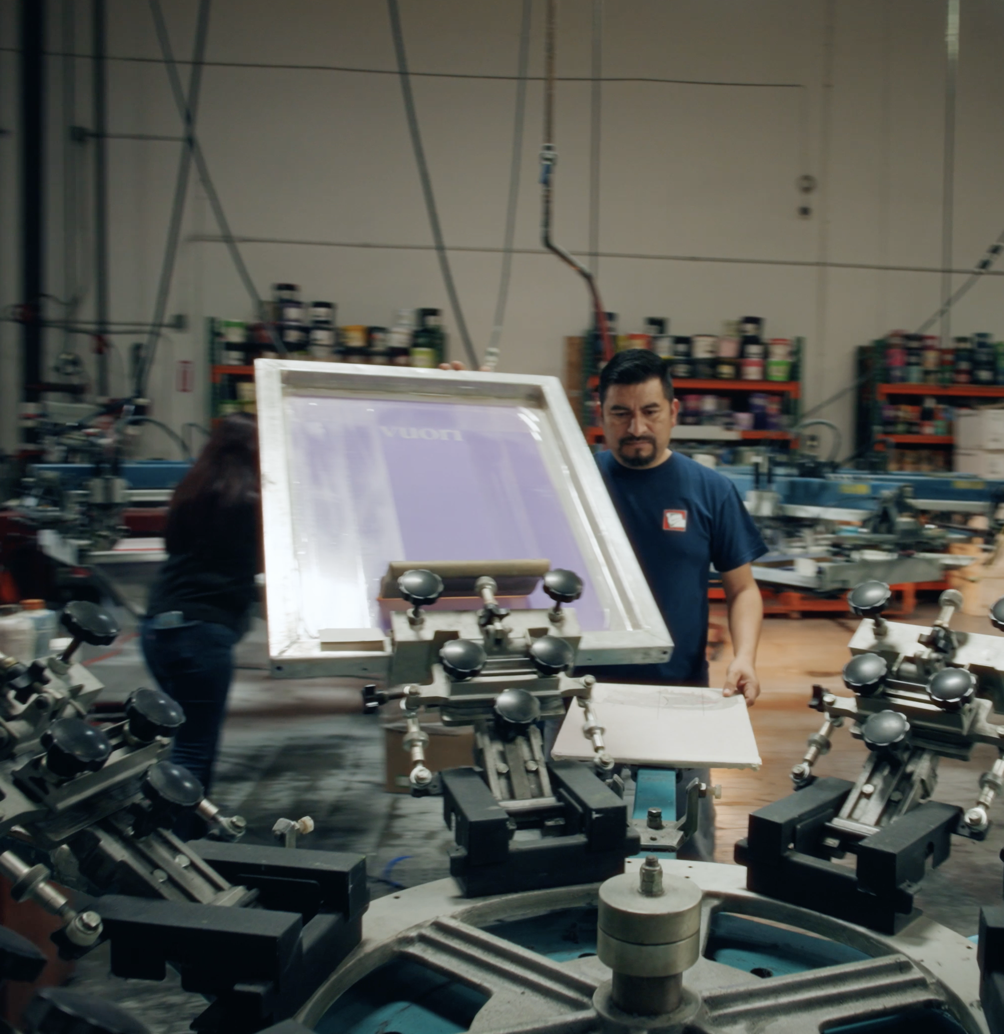 A partially assembled industrial machine with various metallic components, rollers, and belts, set in a factory environment with shelves of tools and supplies in the background.