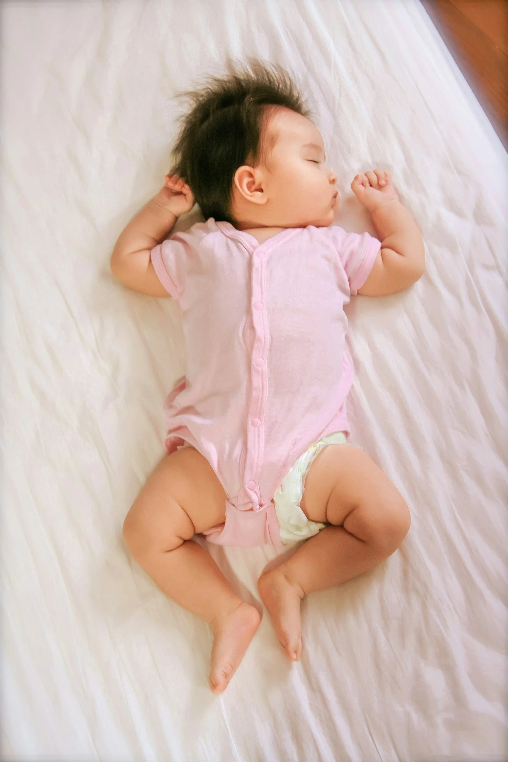 A sleeping baby lying on a white sheet, wearing a pink onesie and a diaper, with curly arms and legs, and dark hair.