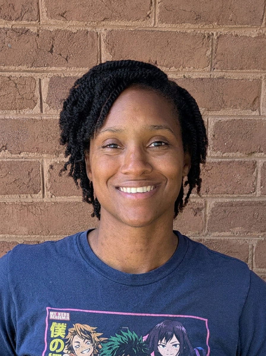 A woman with short black twisted hair smiling in front of a brick wall, wearing a dark blue T-shirt with anime characters printed on it.
