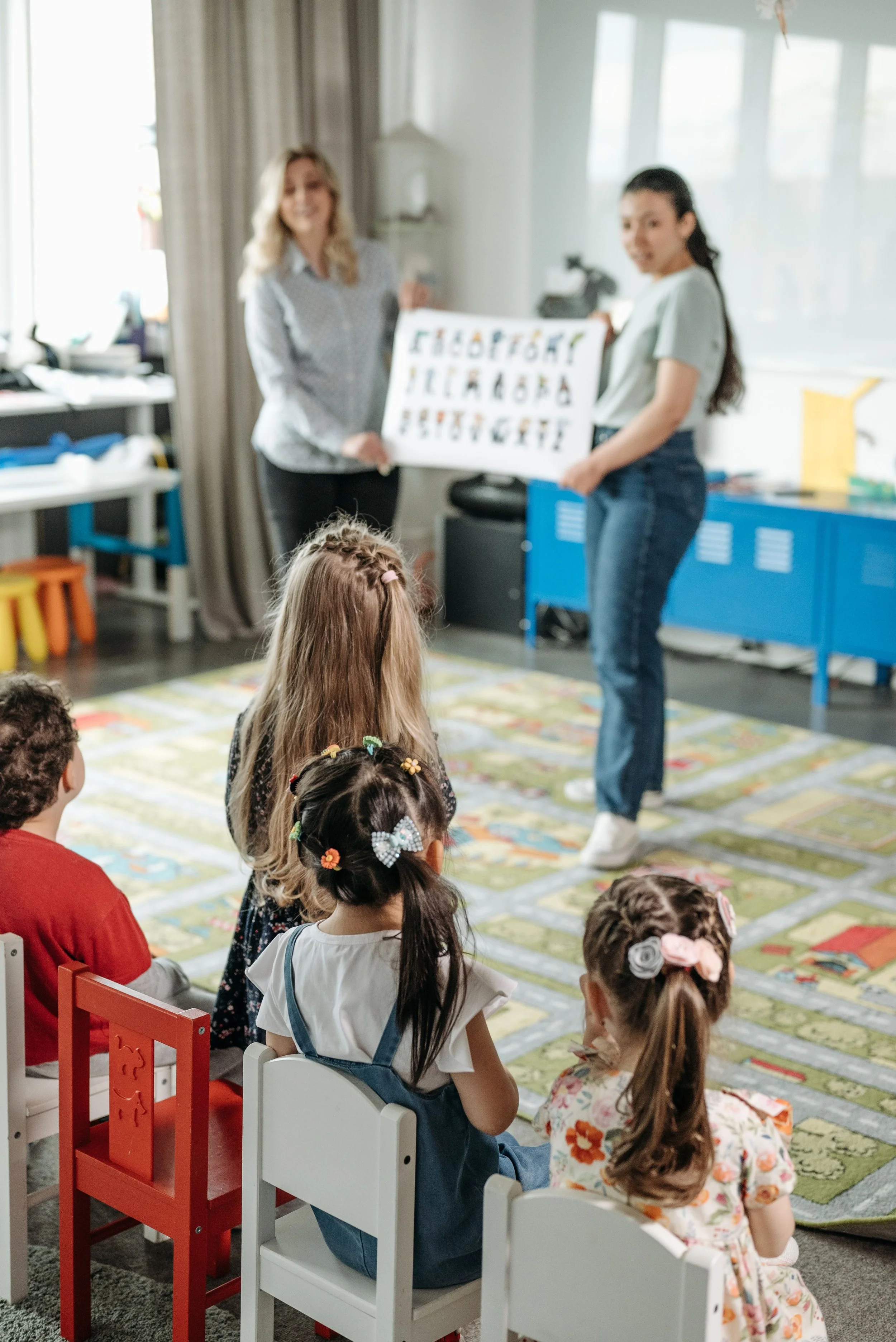 Children seated in a classroom listening to a woman holding an eye chart for vision testing.