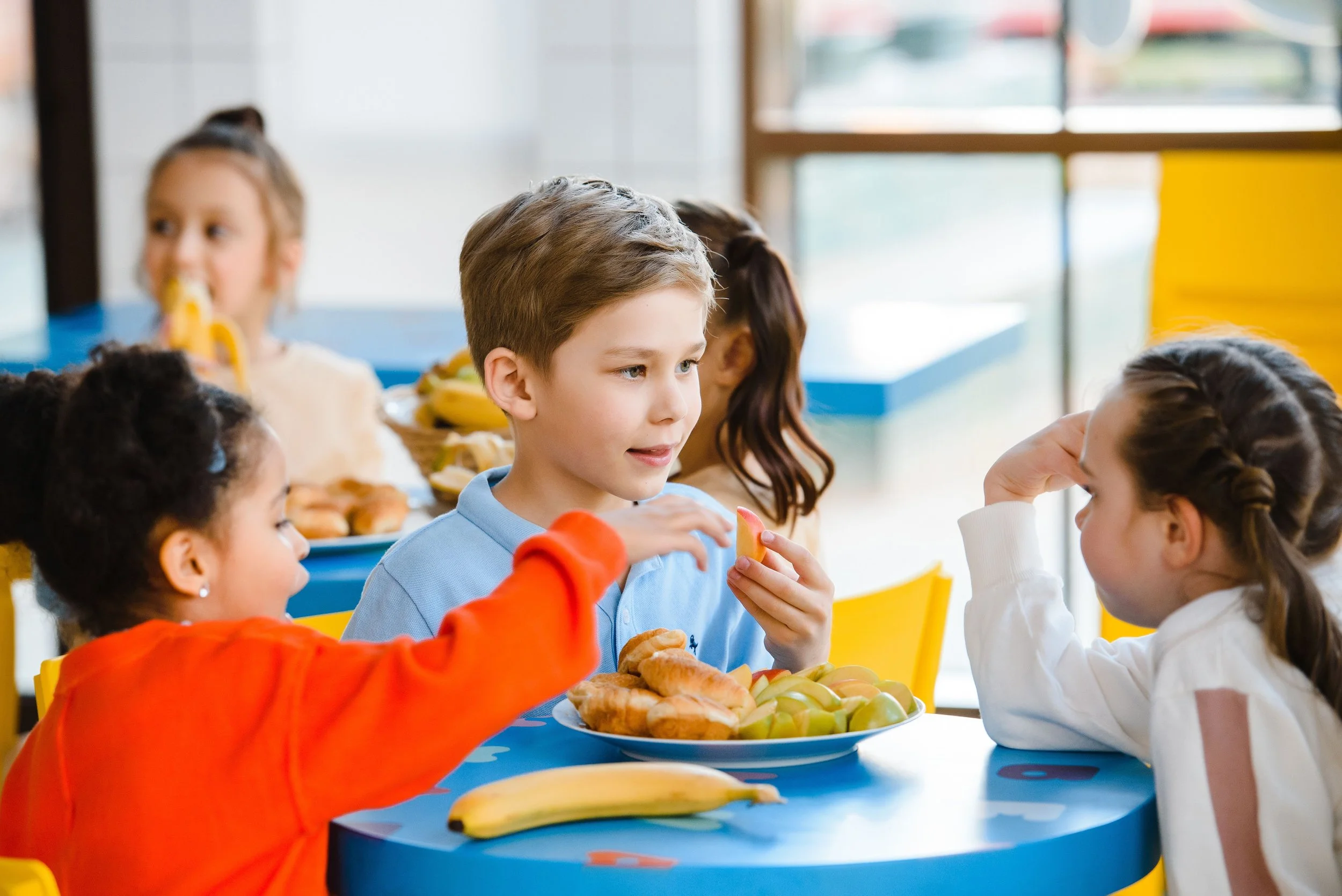 Children sitting at a table with plates of food, including fruits and pastries, inside a bright room with large windows.