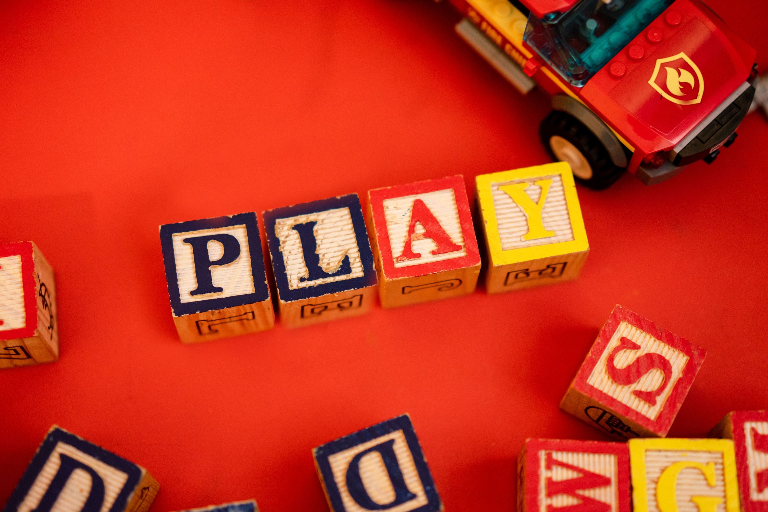 Colorful toy blocks spelling 'PLAY' on a red surface with a toy truck nearby.