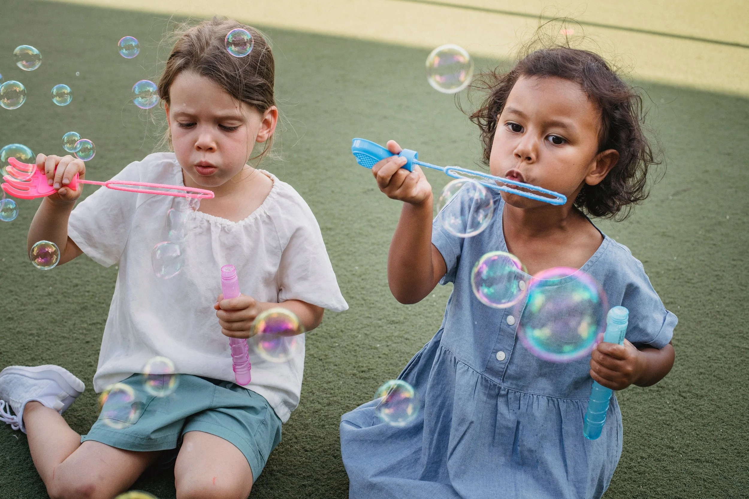 Two young girls sitting on a grassy field blowing soap bubbles with bubble wands.