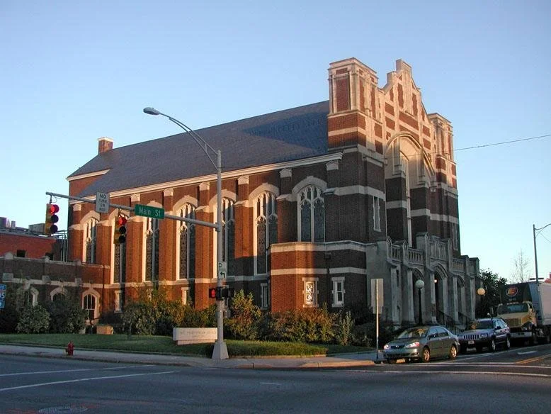 A historic brick church building at the corner of Main Street, with large arched windows, decorative brickwork, and a clock tower. Cars parked along the street and a traffic light at the intersection.
