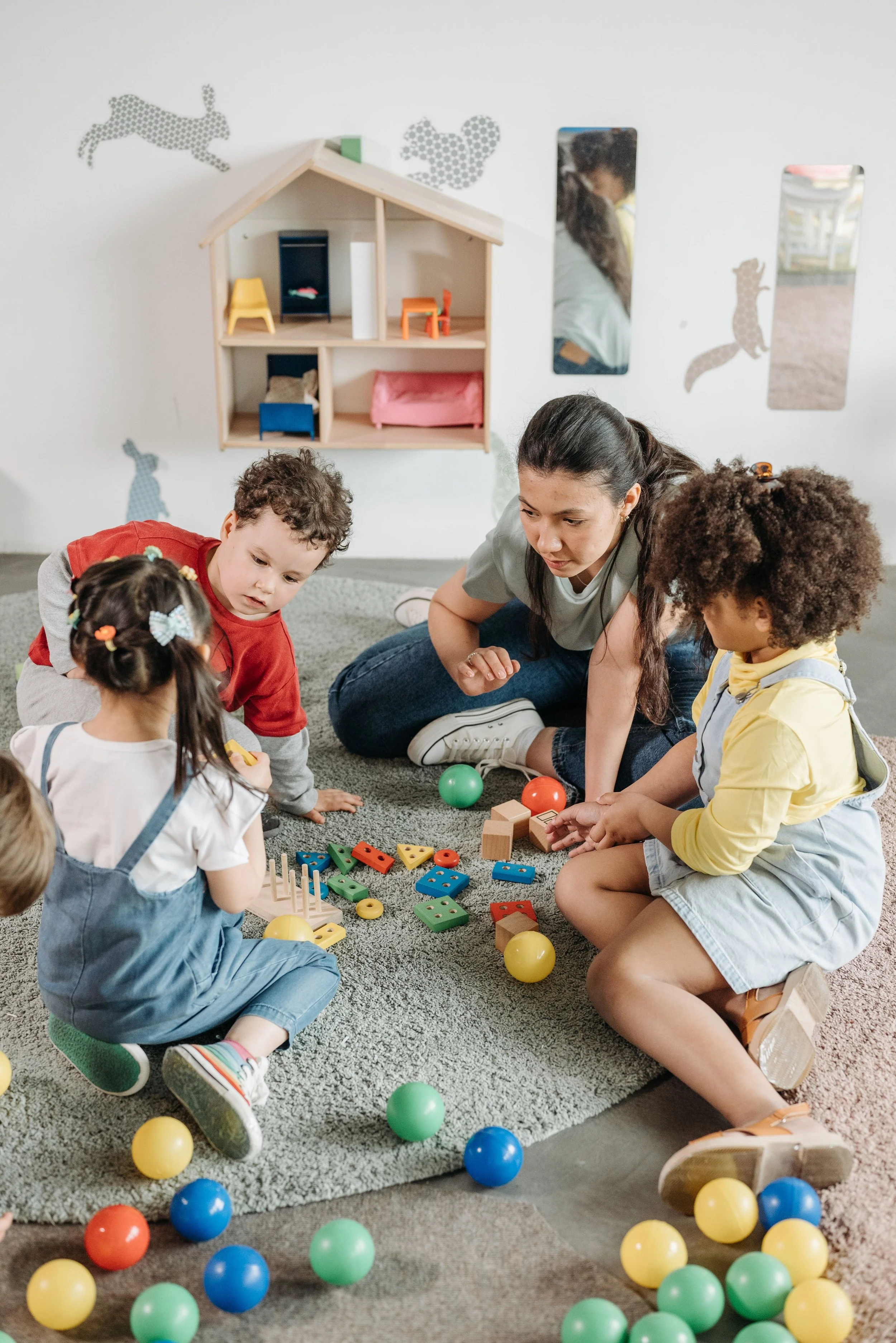 Children and a young woman sitting on a gray carpet playing with colorful educational toys, with scattered plastic balls around them and picture frames on a white wall in the background.