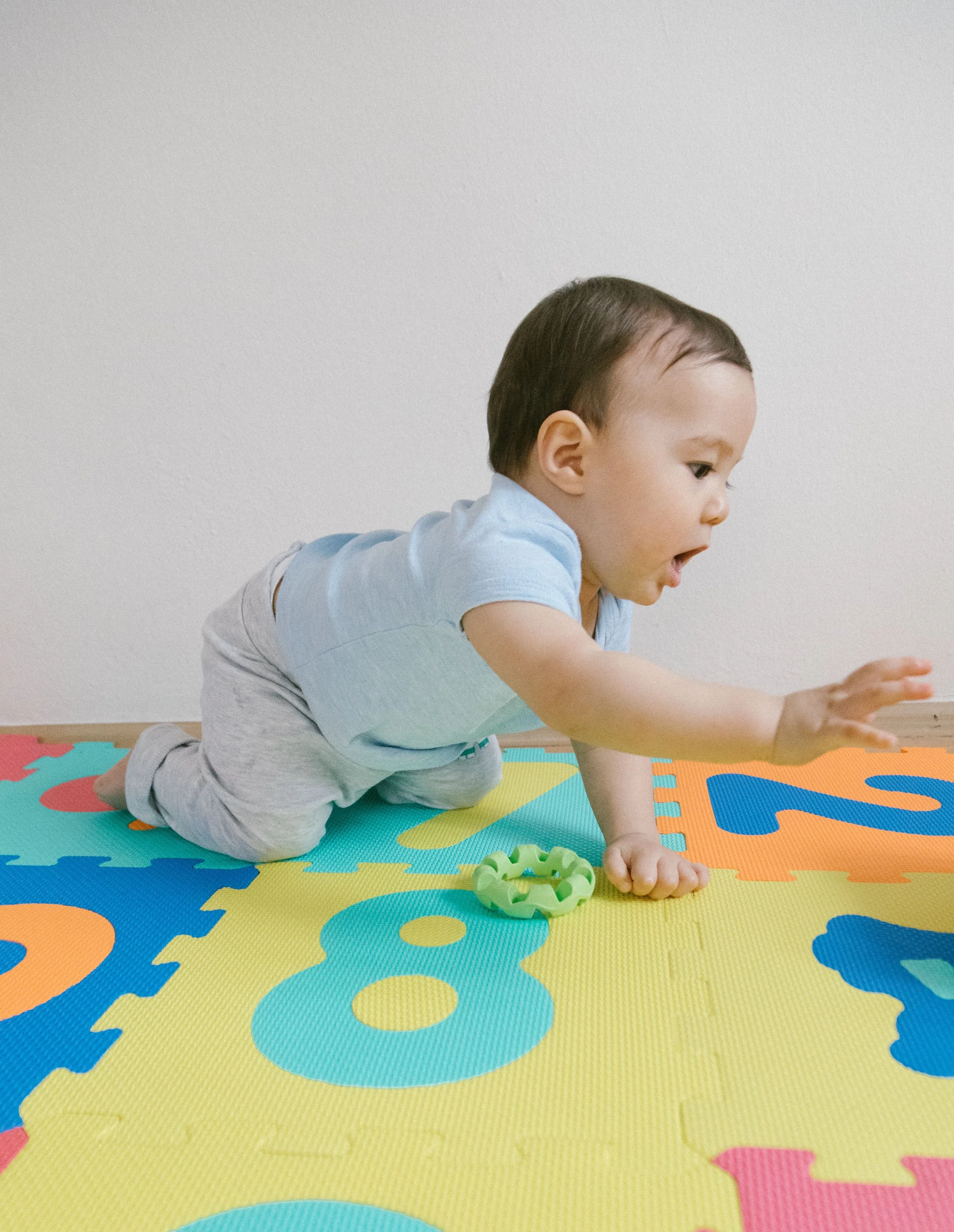 Young boy in a light blue T-shirt and gray pants on a multicolored foam puzzle mat, reaching out with one hand while looking down, with a surprised or excited expression.
