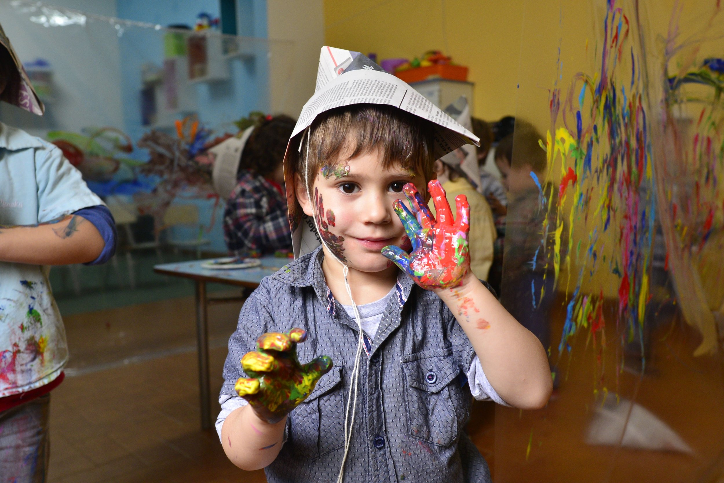 A young boy with messy hair and painted face, wearing a newspaper hat, showing his colorful hands covered in paint in an art classroom.