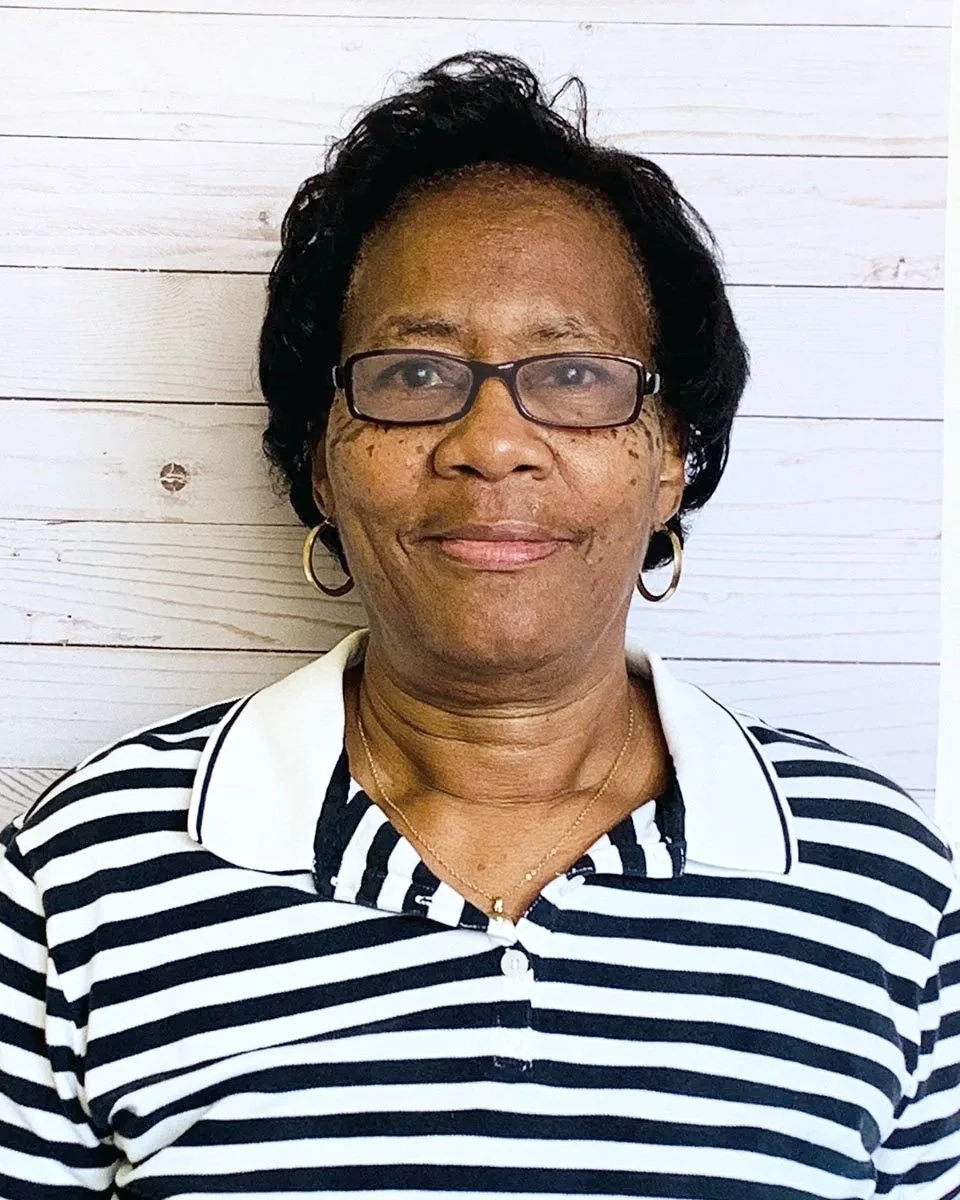 An woman with short black hair, wearing glasses, gold hoop earrings, and a striped black and white polo shirt, standing in front of a white wooden wall.
