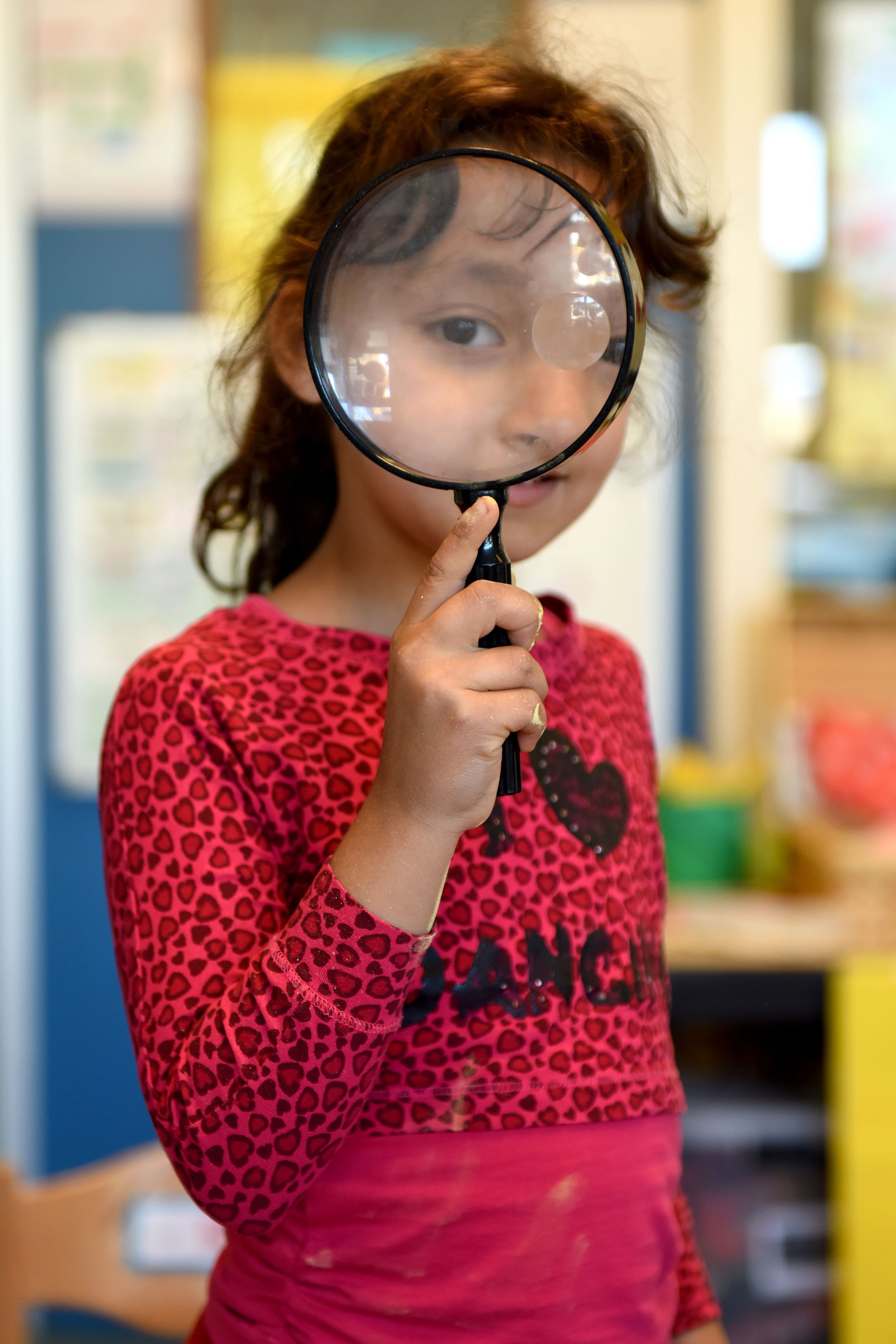 A young girl in a red, pink, and black leopard print shirt holds a magnifying glass in front of her face, partially obscuring it, in an indoor setting.