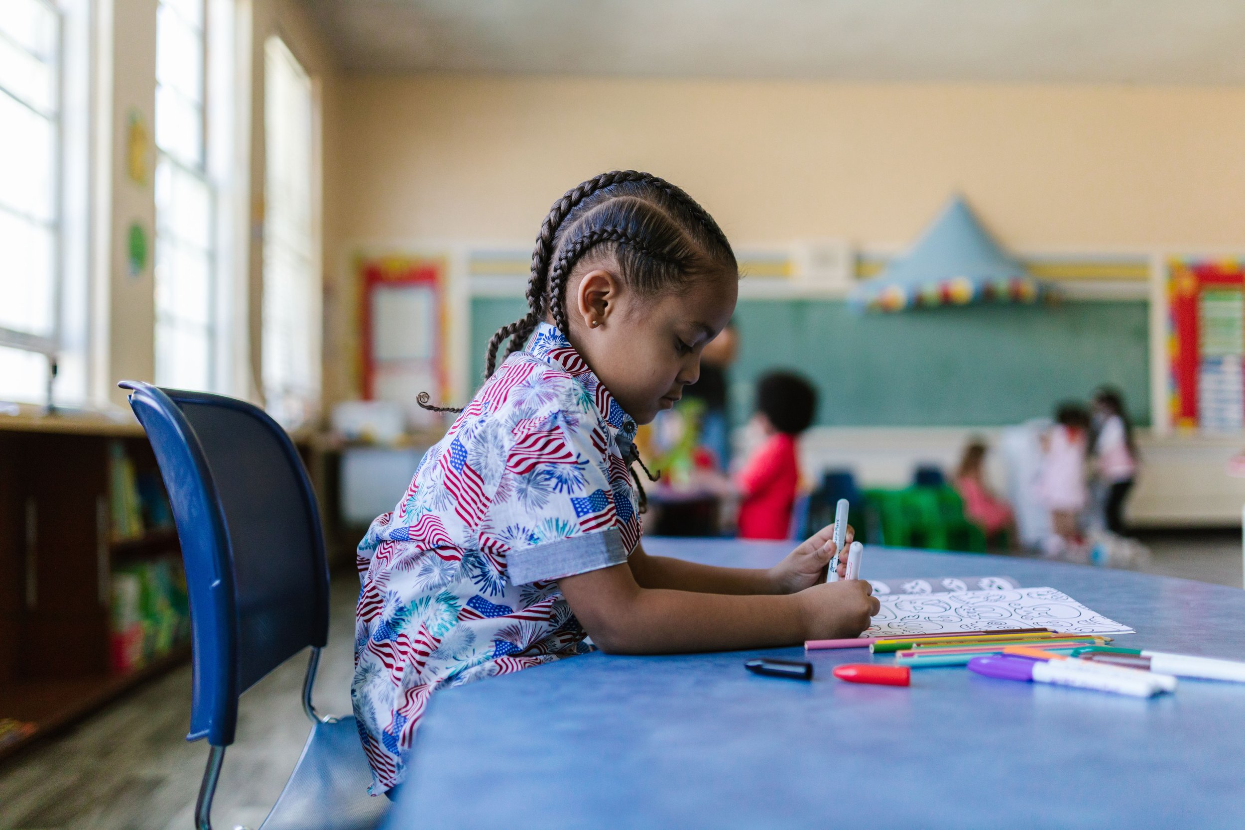 A young girl with braided hair sitting at a table in a classroom, coloring with markers. Other children and classroom decorations are visible in the background.