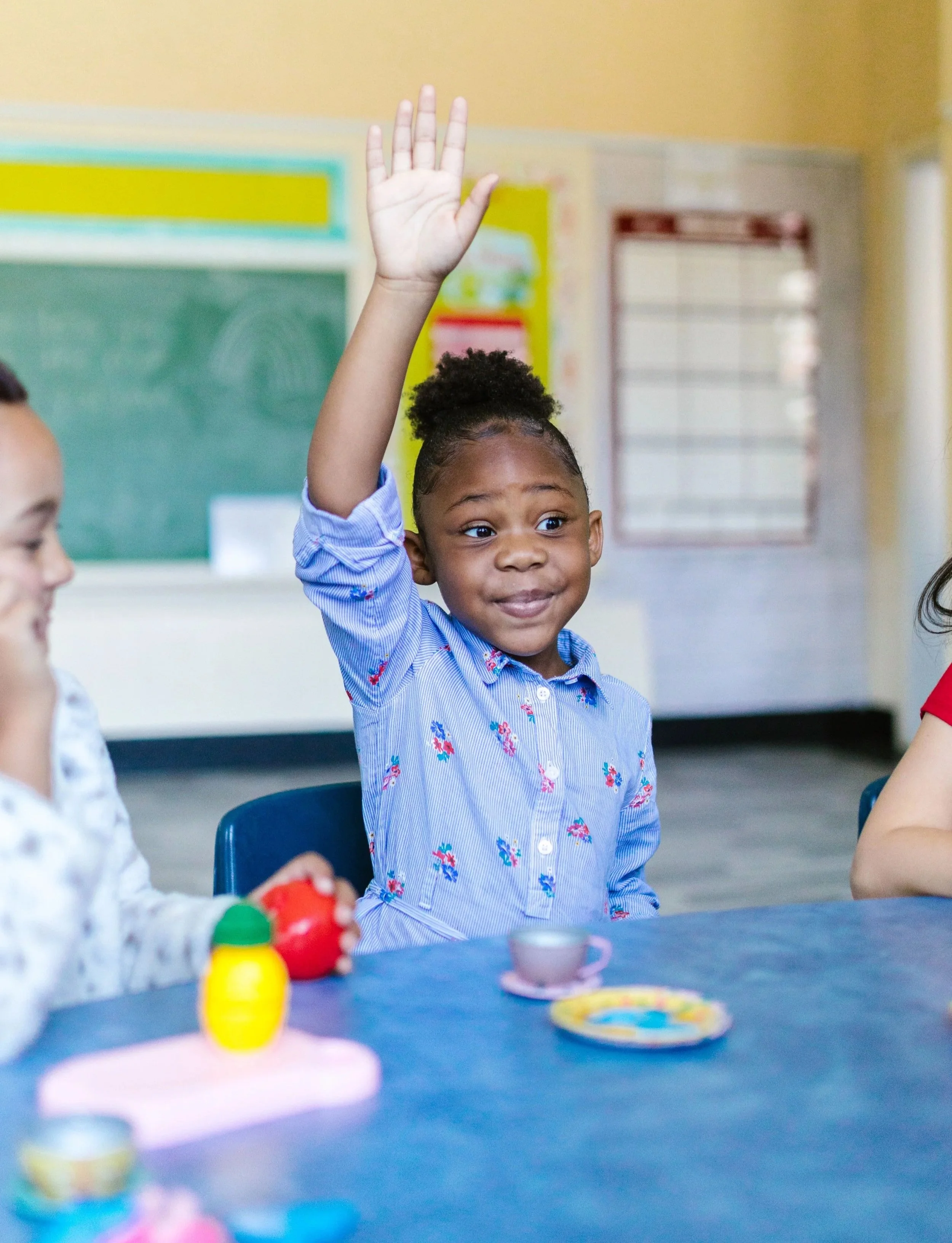 A young girl with dark hair in a puff, wearing a blue patterned shirt, raises her hand in a classroom. Other children are present, and colorful toys and a teacup are on the table. The background includes a green chalkboard and yellow walls.