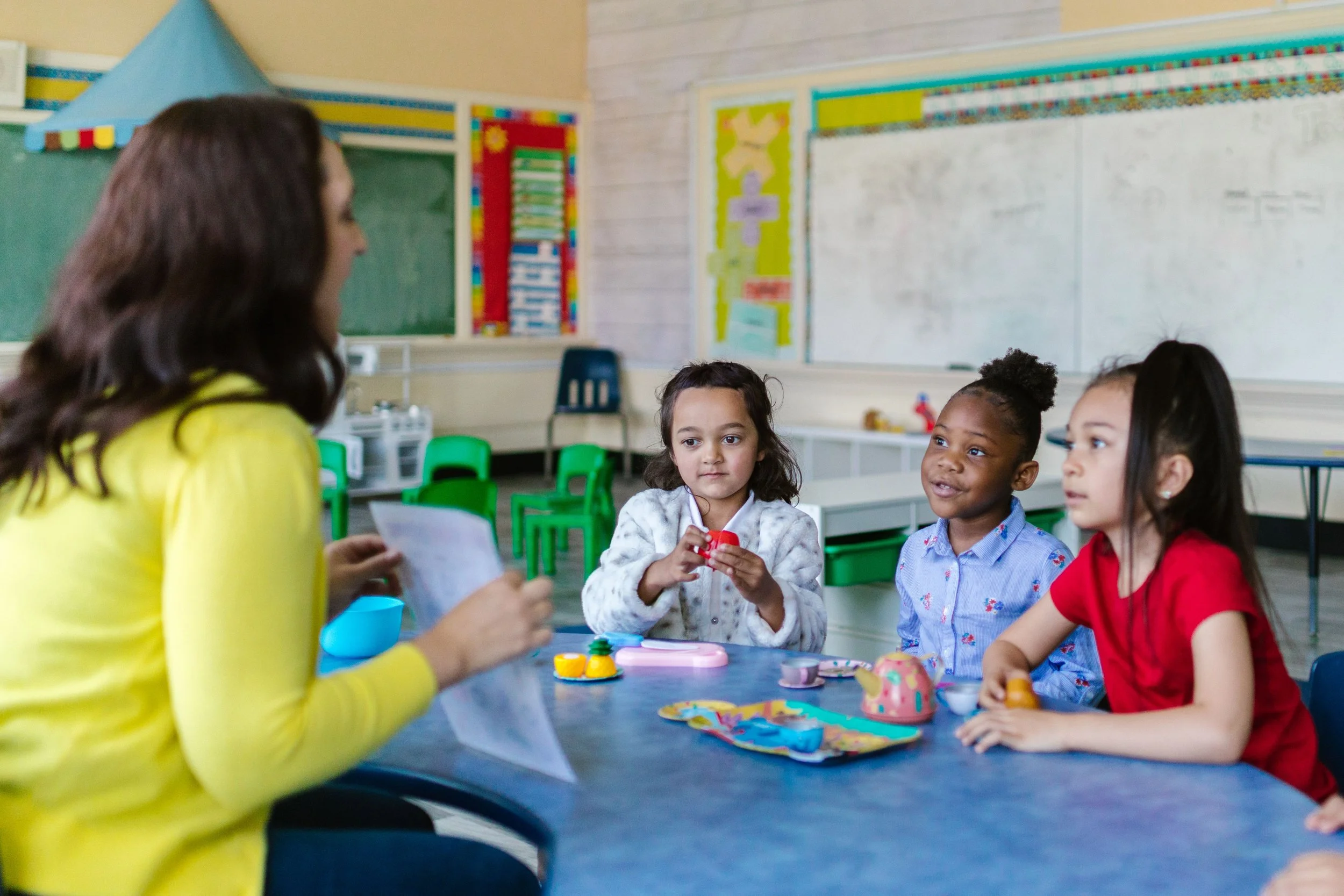 A teacher sitting at a table with four young girls in a classroom. The teacher is wearing a yellow top, and the girls are engaging with toys and looking at the teacher. The classroom has colorful decorations and educational posters on the walls.