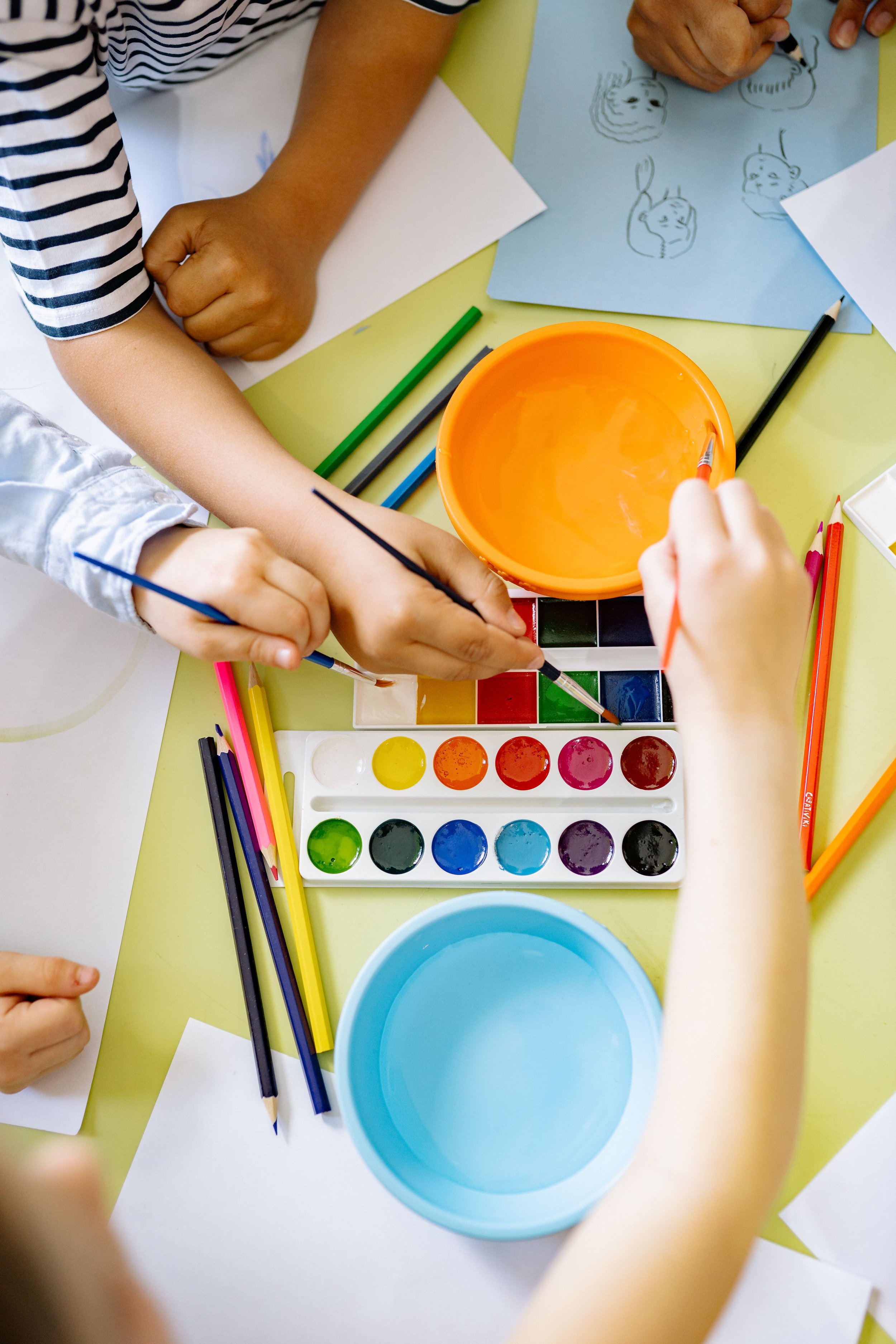 Children drawing and painting with watercolor paints and colored pencils on a table with blank sheets of paper.