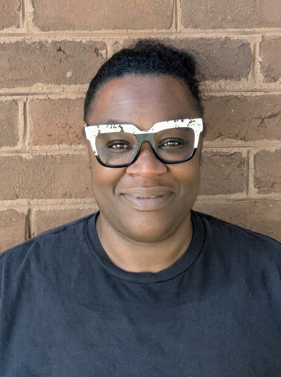 A woman with dark skin, short black hair in a bun, wearing large black and white glasses and a black T-shirt, standing in front of a brick wall.