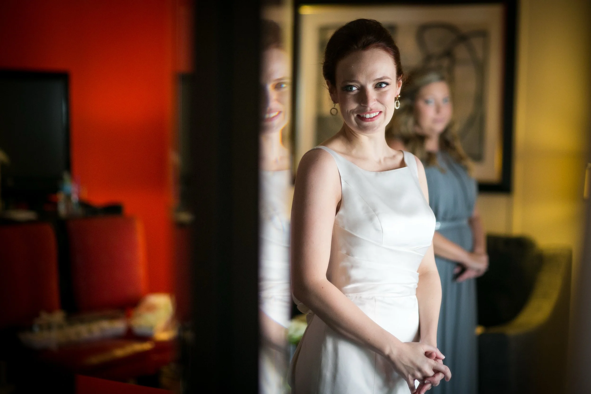 A woman in a white dress stands in a room, with another woman in a gray dress in the background. The room has colorful walls and framed artwork. The woman in white is smiling and appears to be in a joyful moment.
