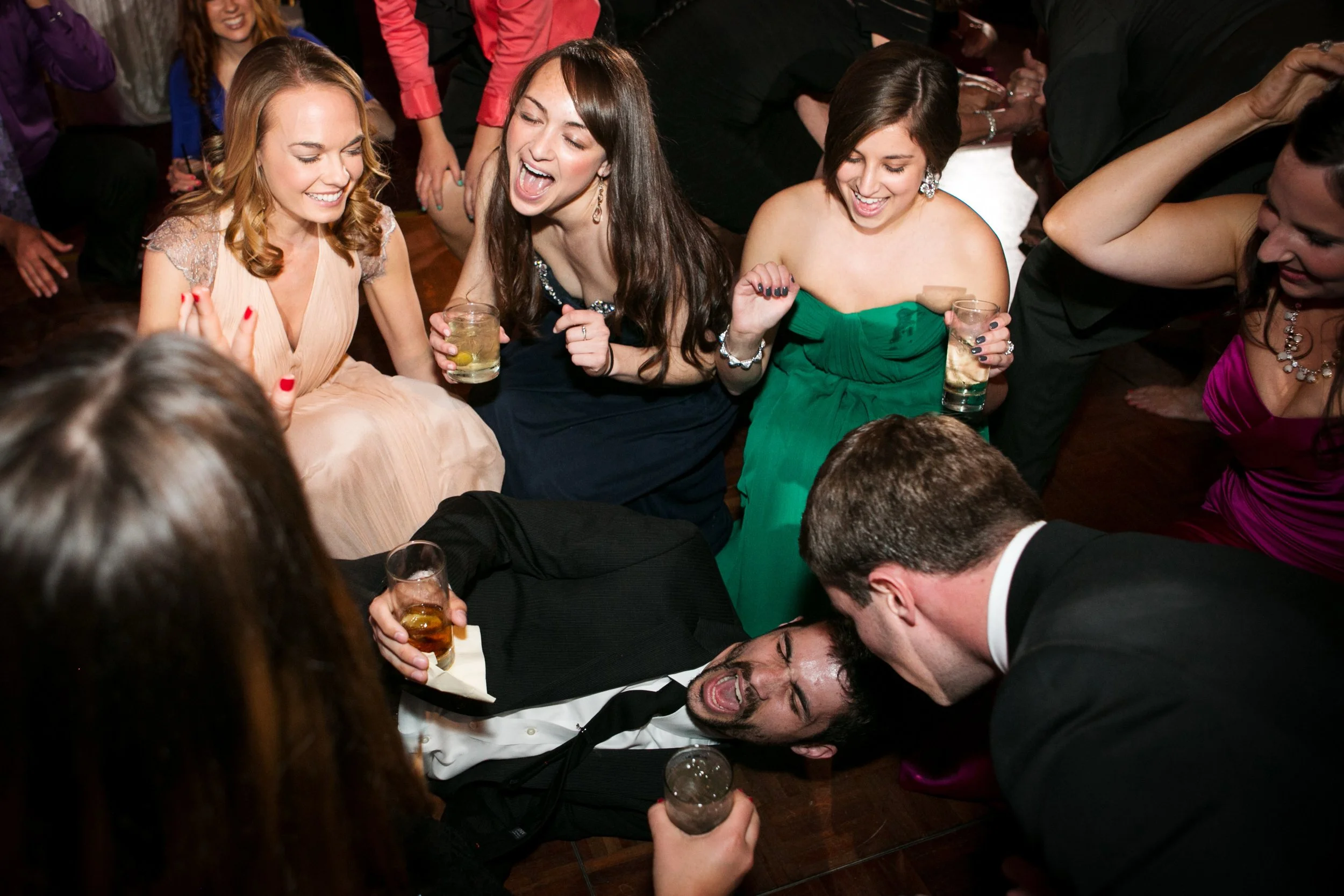 Group of young adults dancing and partying at a celebration with drinks, some sitting and some standing, lively and fun atmosphere.